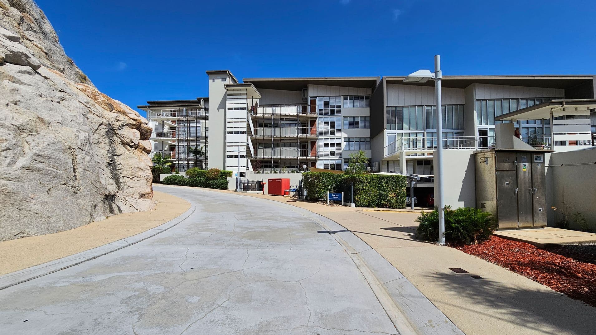 A modern building with large windows and balconies is on the right, surrounded by greenery and a paved walkway. A rocky cliff is on the left, with a smooth road curving between the cliff and the building. A streetlamp and some shrubs are in the foreground, and a small red utility box is visible near the building. The sky is clear and blue.