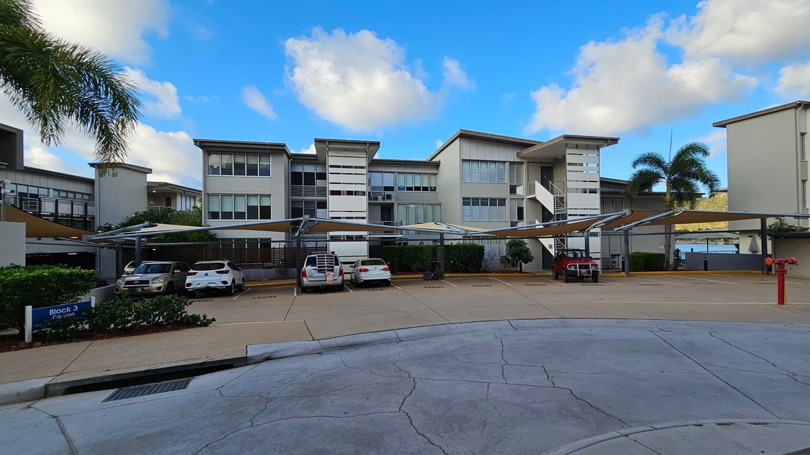 A modern apartment complex with three-storey buildings in the background. In the foreground, there is a car park with several vehicles under a metal canopy. A sign reads "Block 3 Car Park" on the left. Palm trees are visible on both sides, and a body of water can be seen in the distance on the right. The ground is paved with concrete, and there is a red fire hydrant on the right.