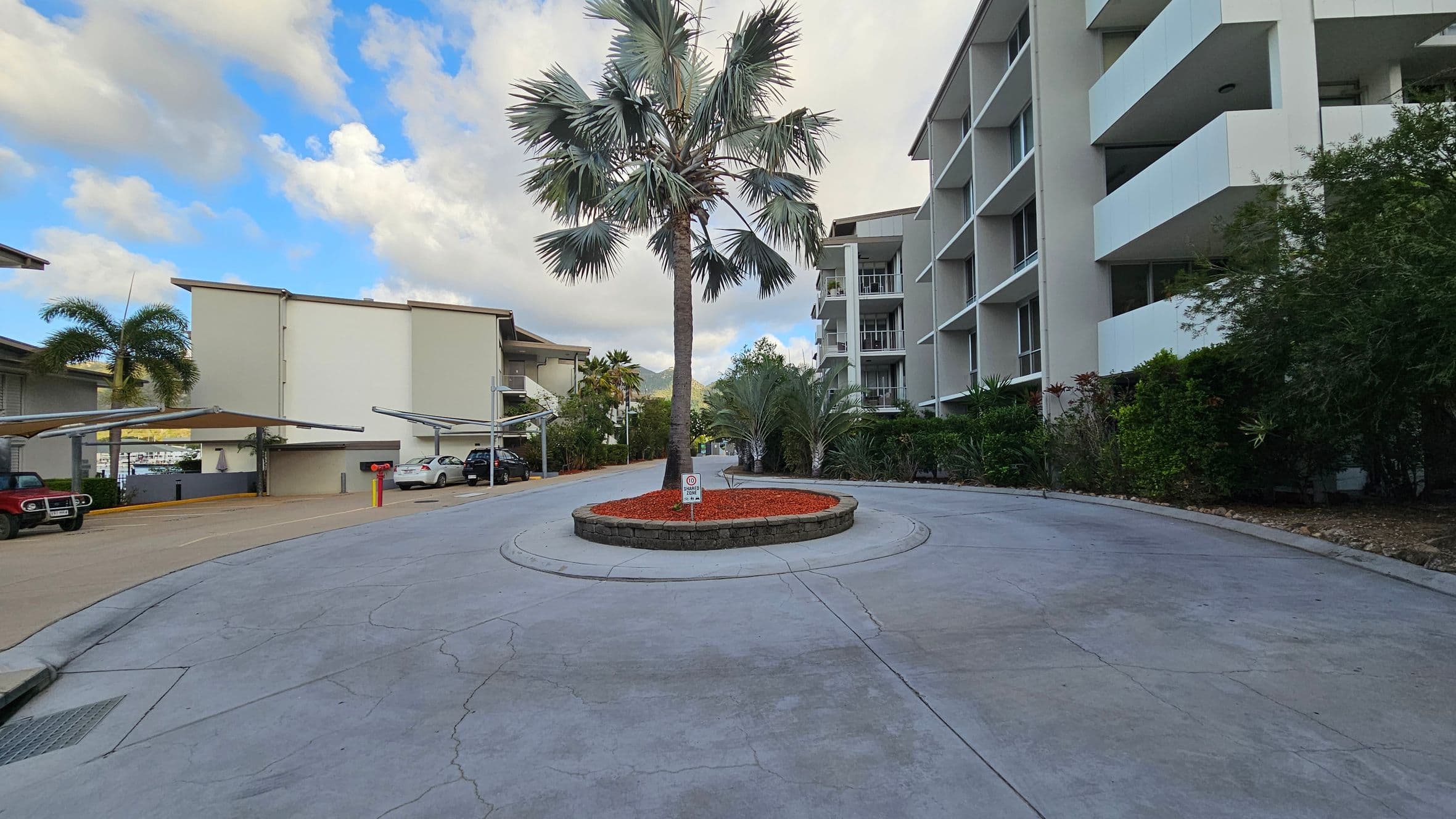 A roundabout with a tall palm tree in the centre, surrounded by red mulch. On the right, a multi-storey building with balconies and greenery. On the left, a covered parking area with several cars, including a red vehicle. The ground is paved with visible cracks, and there are various plants and trees around the area.
