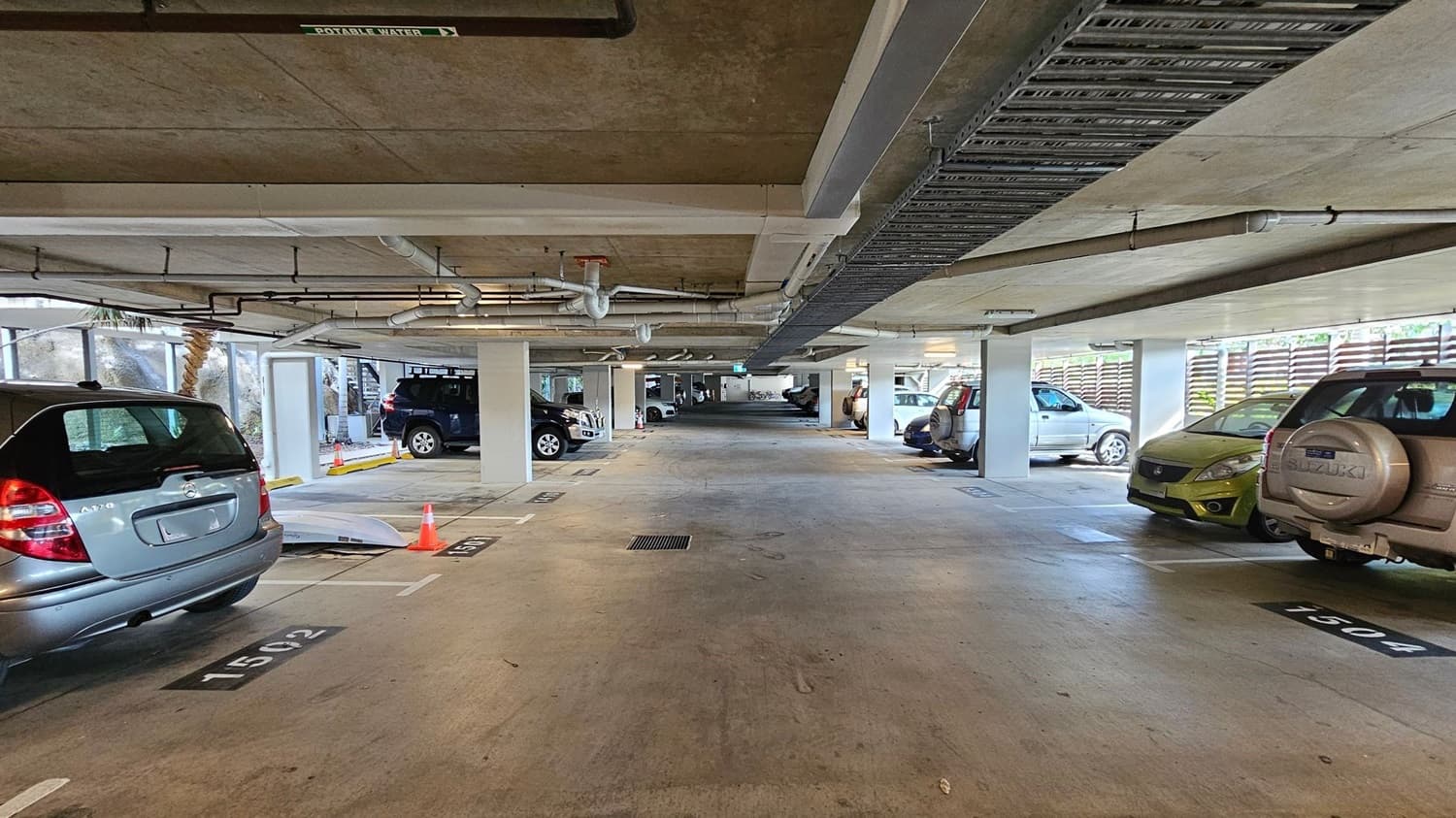 A covered car park with several vehicles parked on both sides. On the left, a silver car with a visible number plate is parked in space 1502. On the right, a beige SUV is parked in space 1504. The floor is concrete, and the ceiling has exposed pipes and a sign indicating "Potable Water." Orange traffic cones are placed near the entrance. The background shows more parked cars and a bicycle.
