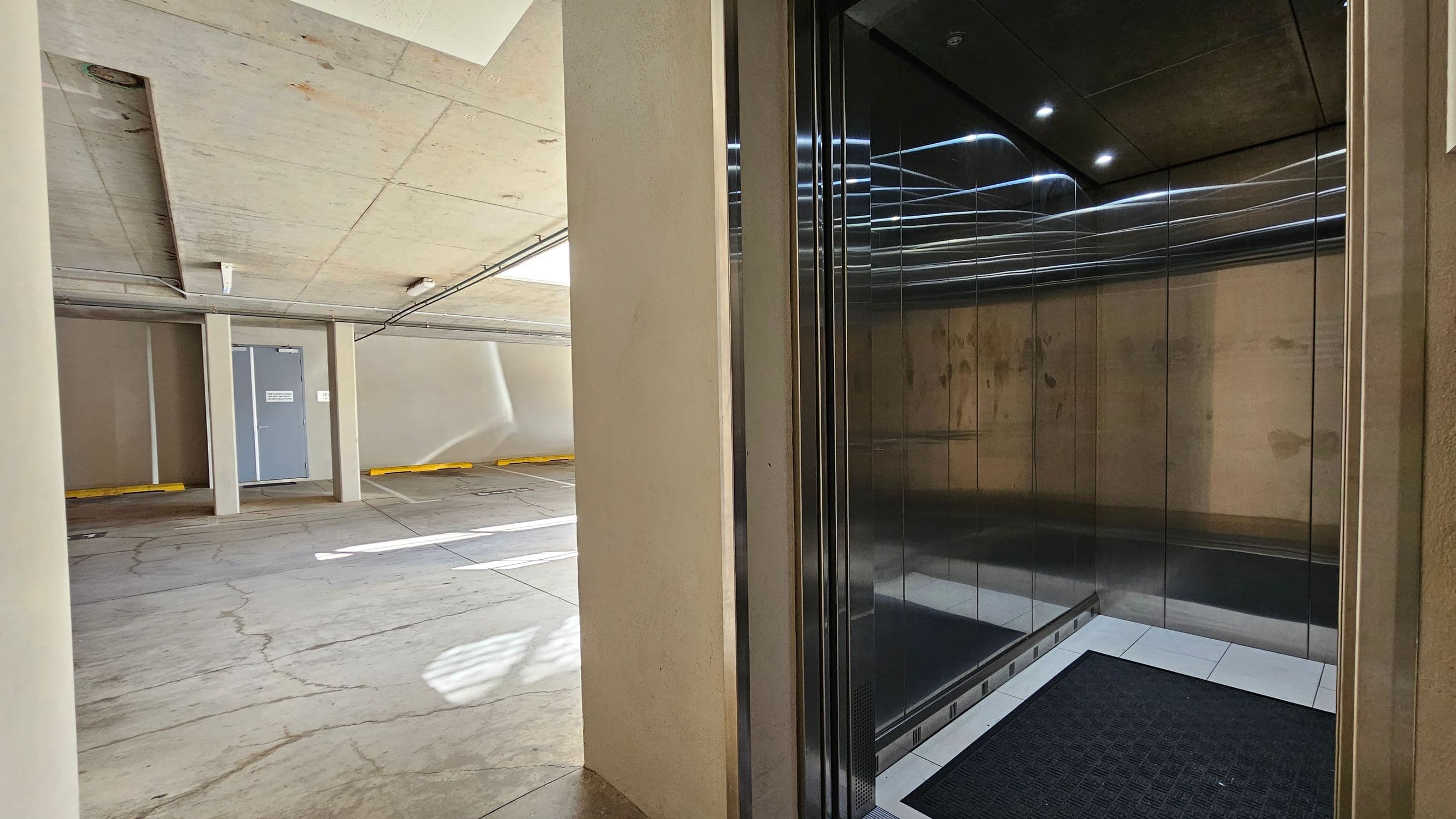 On the left, a concrete parking area with a grey door and yellow parking stops. The floor is grey with visible cracks. The ceiling is concrete with a light fixture. On the right, an open lift with reflective metal walls and a black floor mat. The lift interior is illuminated by small ceiling lights.