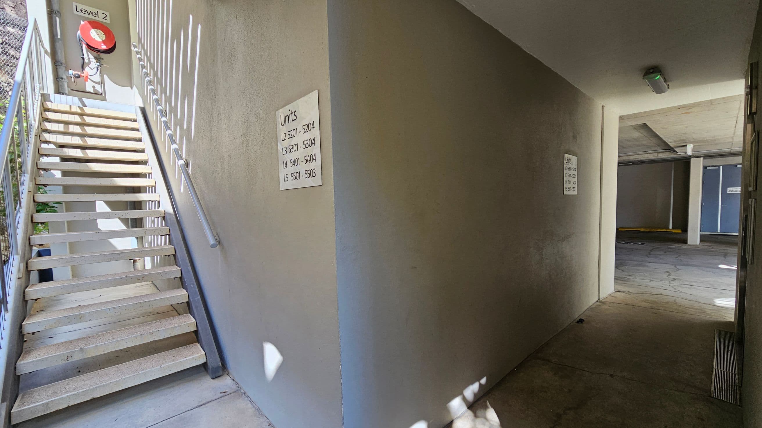A stairway on the left with metal railings leads up to a landing marked "Level 2" with a red fire hose reel. On the wall, a sign lists unit numbers for levels 2 to 5. To the right, a corridor leads to a dimly lit parking area with concrete flooring and a ceiling light. The parking area has blue doors and structural columns. Shadows and light patterns are visible on the walls and floor.