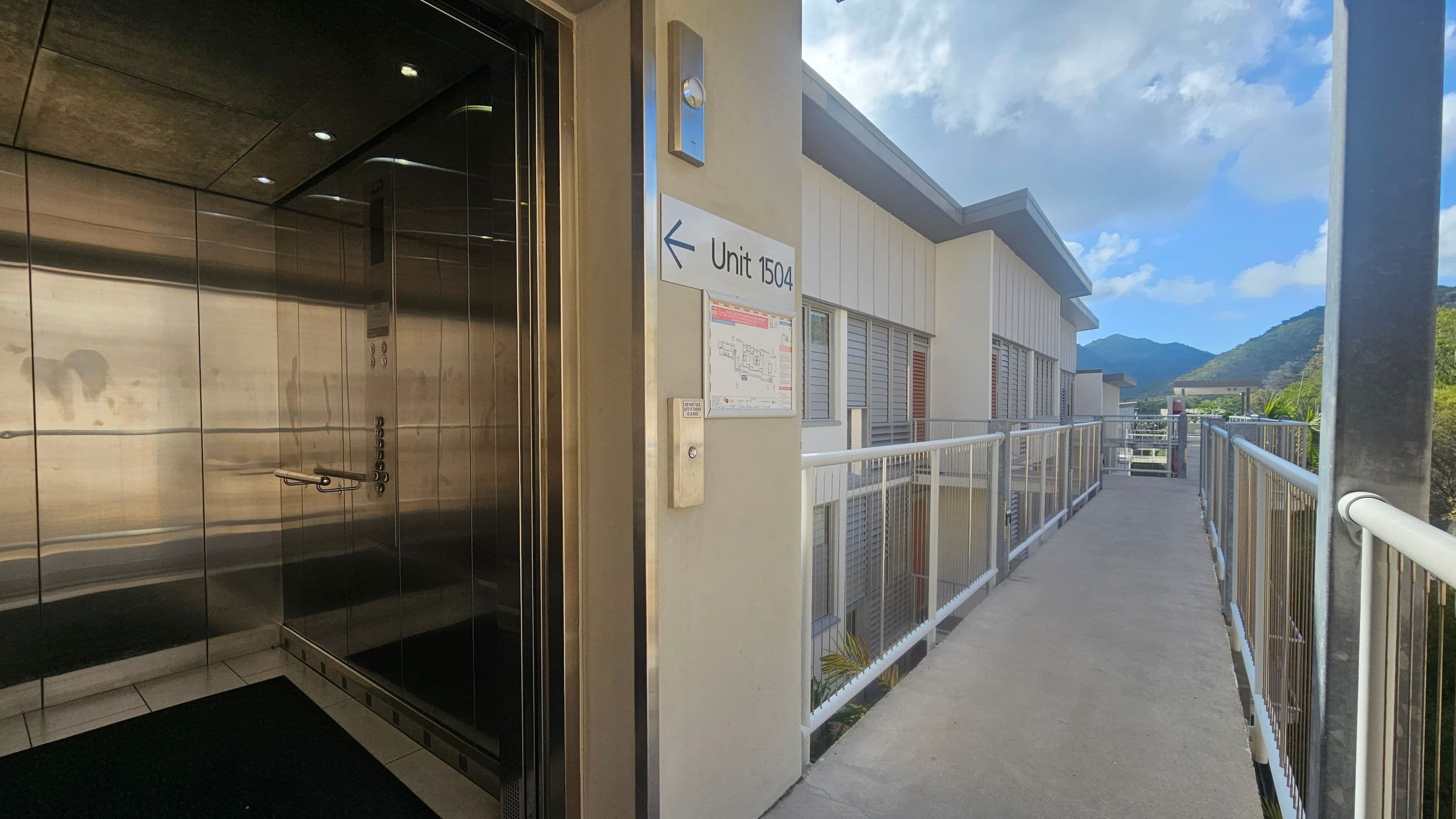 On the left, an open lift with metallic walls and a black floor. In the middle, a wall with a sign pointing to "Unit 1504" and a map below it. On the right, a walkway with metal railings leading to a building with louvred windows. In the background, there are green hills and a partly cloudy sky.