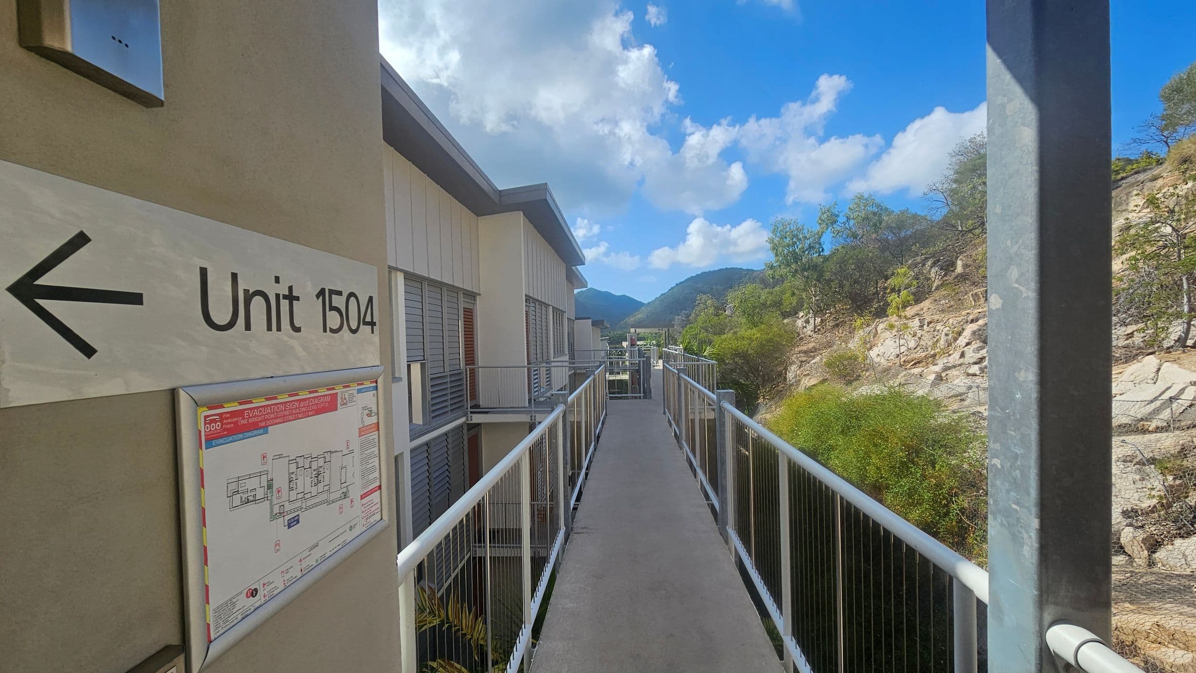 A walkway with metal railings leads towards a building with louvred windows on the left. A sign on the left wall reads "Unit 1504" with an arrow pointing left, and below it is an evacuation diagram. On the right, there is a rocky hillside with sparse vegetation. In the background, there are mountains under a partly cloudy sky.