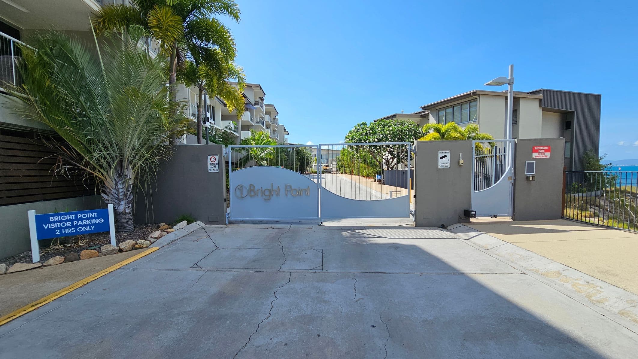 A paved area with visible cracks leads to a gated entrance on the left, marked "Bright Point". Behind the gate, a building is partially visible, surrounded by greenery including palm trees. On the right, a metal fence runs parallel to the road, overlooking a body of water and distant hills. The sky is clear with some clouds. Yellow bollards are in the foreground near the fence.