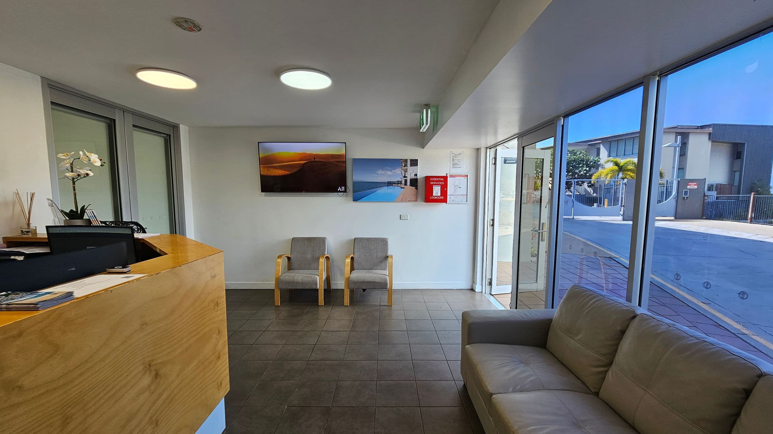 A reception area with a wooden desk on the left, featuring a computer and a potted orchid. Two grey chairs are in the centre against the wall, with two landscape photographs above them. On the right, a beige sofa is near large glass windows and doors, offering a view of a building and palm trees outside. The floor is tiled, and the ceiling has circular lights.