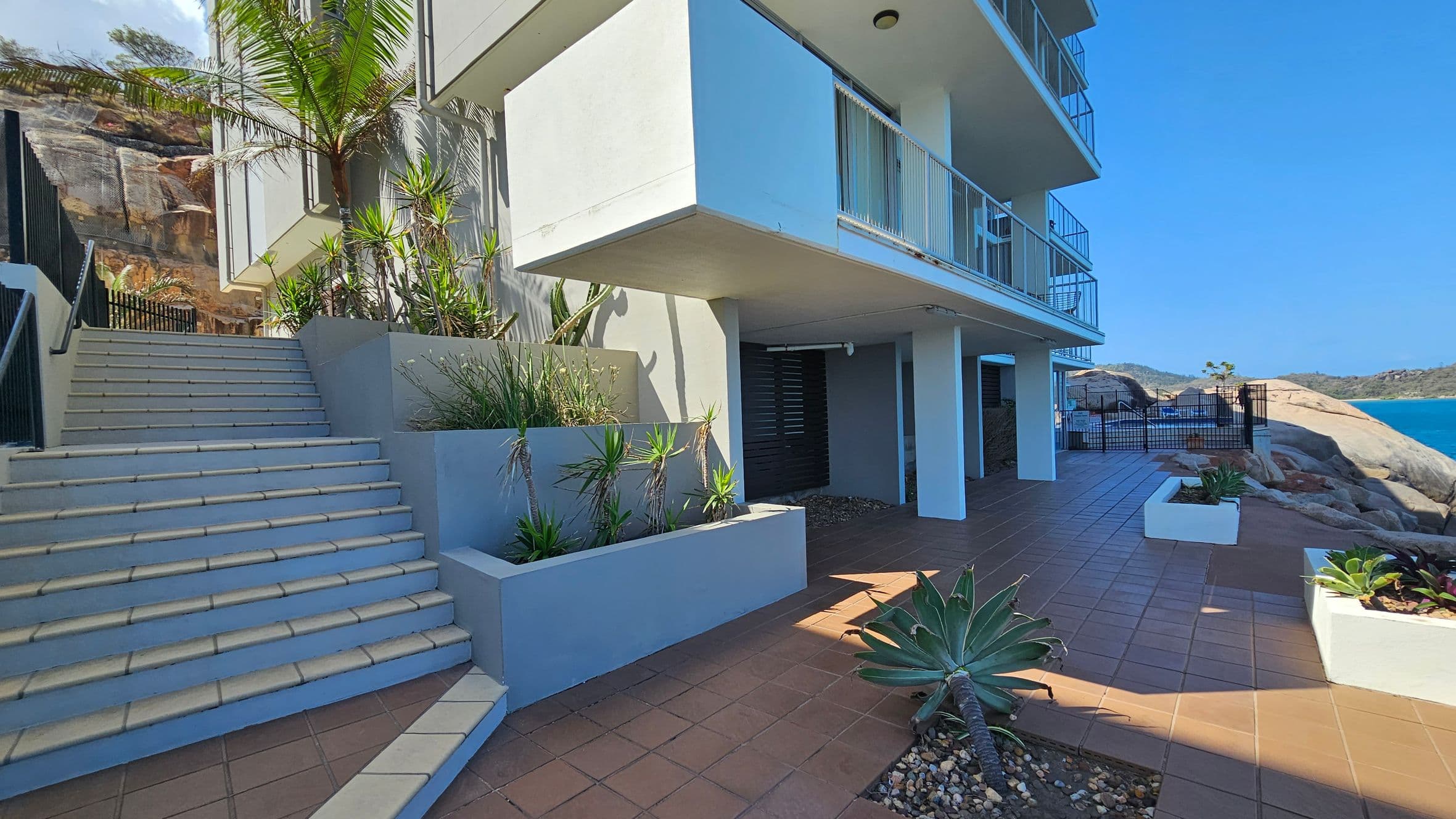 A modern building with a white exterior and balconies on the right. In the foreground, a tiled patio with potted plants and a large agave. On the left, a staircase with light-coloured tiles leads upwards, bordered by planters with tall, spiky plants. In the background, rocky terrain and a view of the ocean with a clear blue sky.