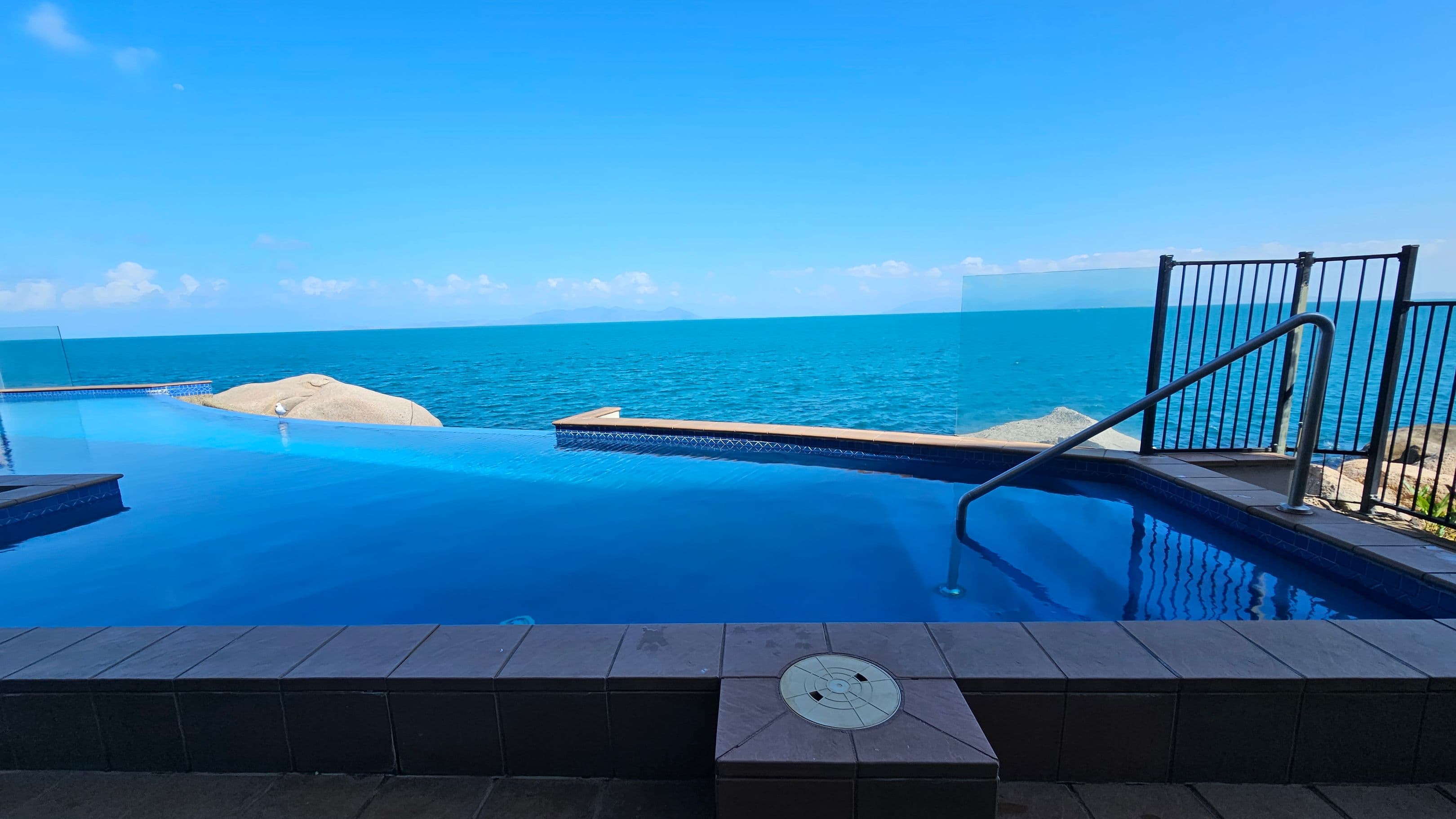 An infinity pool with clear blue water is in the foreground, bordered by dark tiles. A metal handrail descends into the pool on the right. Beyond the pool, the ocean stretches to the horizon, with a rocky outcrop visible on the left. A glass and metal fence runs along the right side, separating the pool area from the sea view.