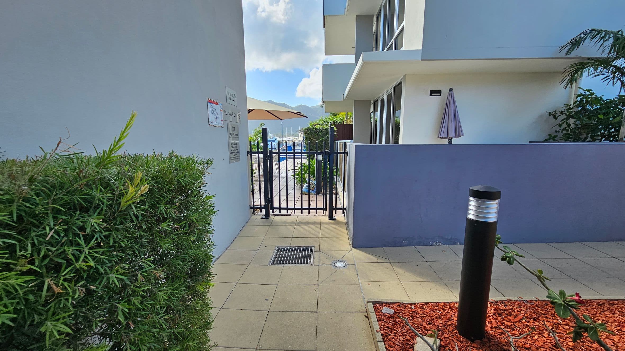 A narrow pathway with tiled flooring leads to a black metal gate. On the left, there's a green hedge and a wall with signs. On the right, a blue wall with a closed umbrella mounted on it. Beyond the gate, a pool area with a large umbrella and chairs is visible. A black bollard light stands on the right, next to a patch of red mulch with a few plants.