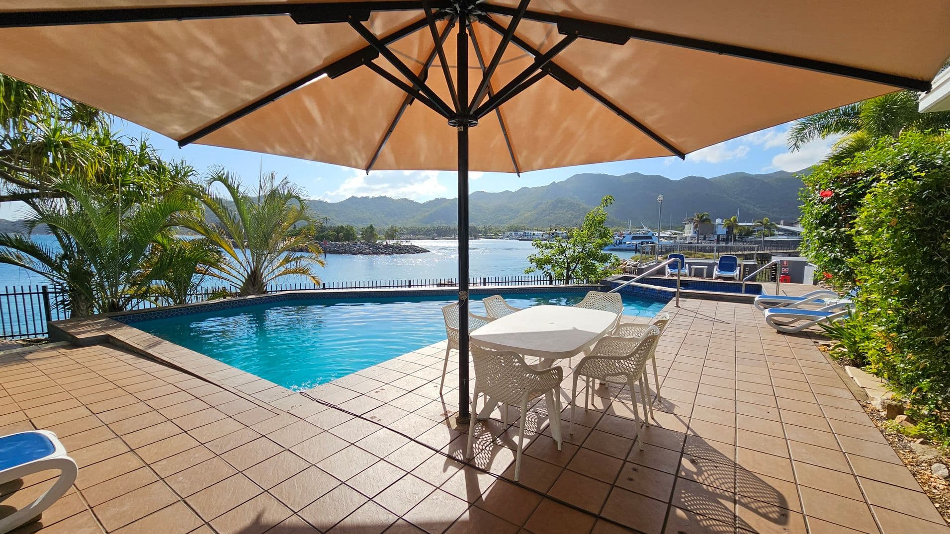 A poolside area with a tiled floor and a large umbrella providing shade over a white table and chairs. The pool is adjacent to a body of water, with a metal fence separating them. Palm trees and lush greenery are on the left and right. In the background, there are mountains and a marina with boats. Two sun loungers are on the right side near the pool.