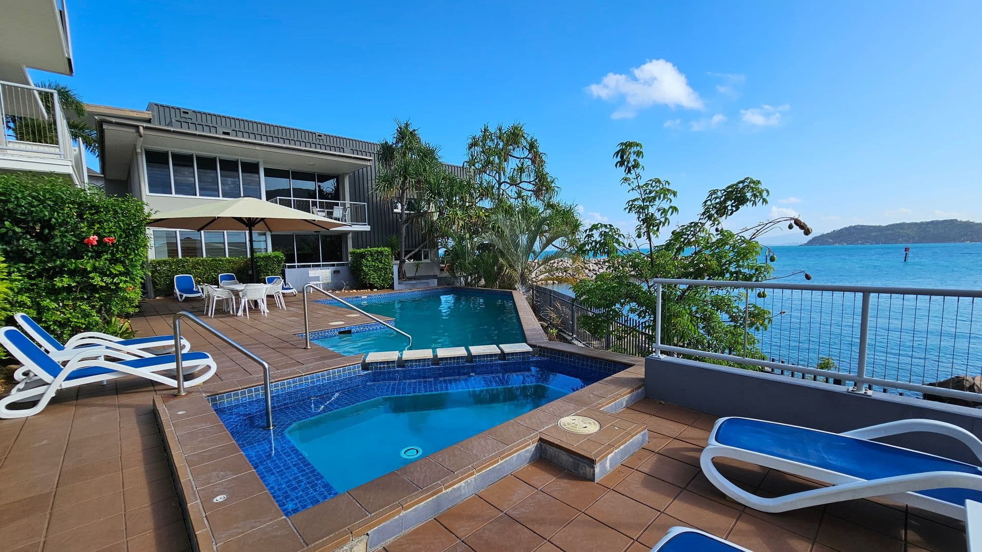 A poolside area with a small hot tub in the foreground, surrounded by brown tiles. Blue and white sun loungers are placed on the left. In the middle, a larger pool is visible, with a few white tables and chairs under a large umbrella. The background features a modern building with large windows and lush greenery, including palm trees. To the right, a railing overlooks a body of water with a distant landmass.