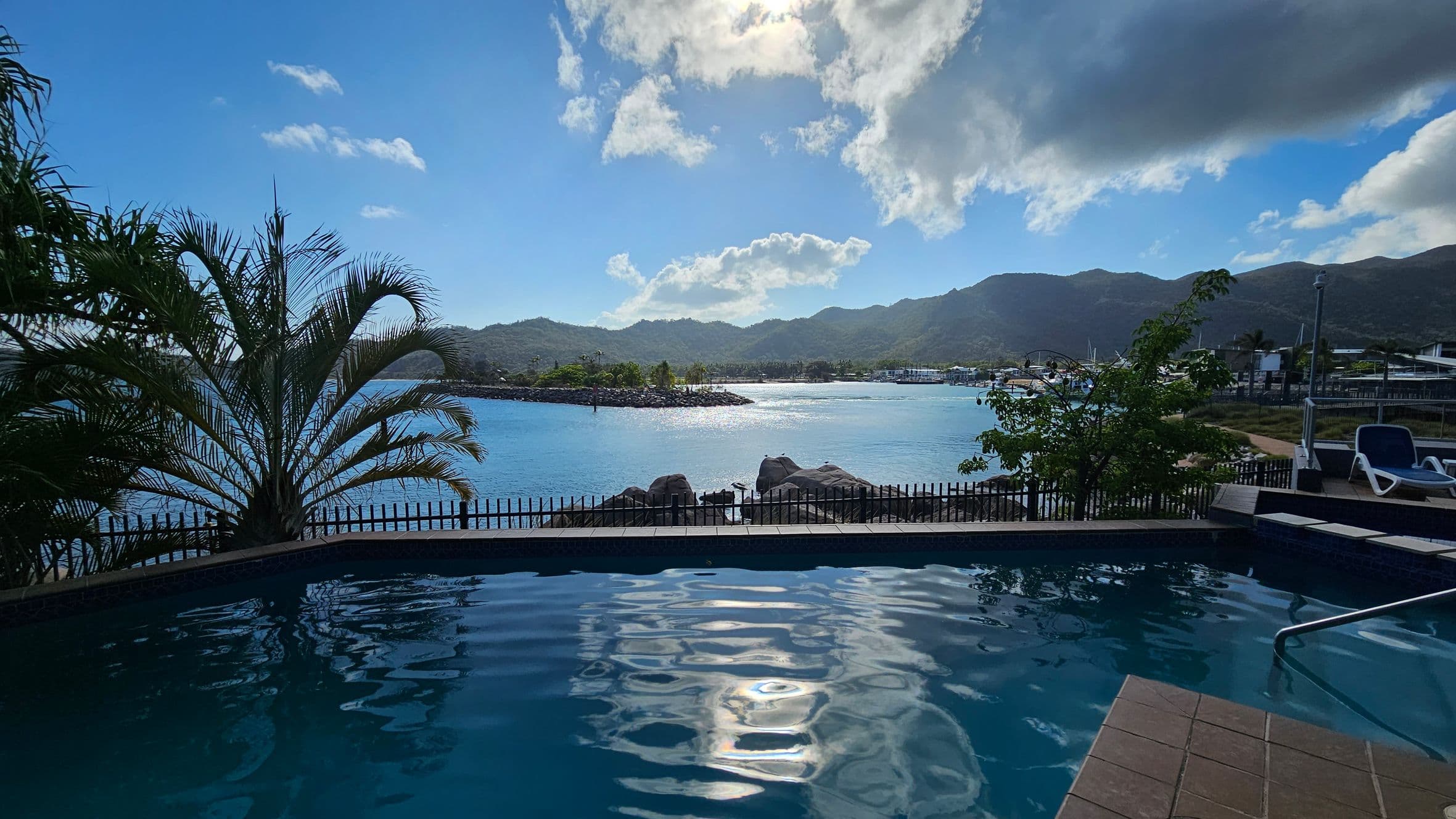 A swimming pool in the foreground with rippling water reflecting sunlight. To the left, a large palm tree is visible, and to the right, a couple of sun loungers are placed near the pool. In the background, a body of water stretches out towards a mountainous landscape. A fence runs along the edge of the pool area, and a few trees are scattered around. The sky is partly cloudy, with the sun peeking through.