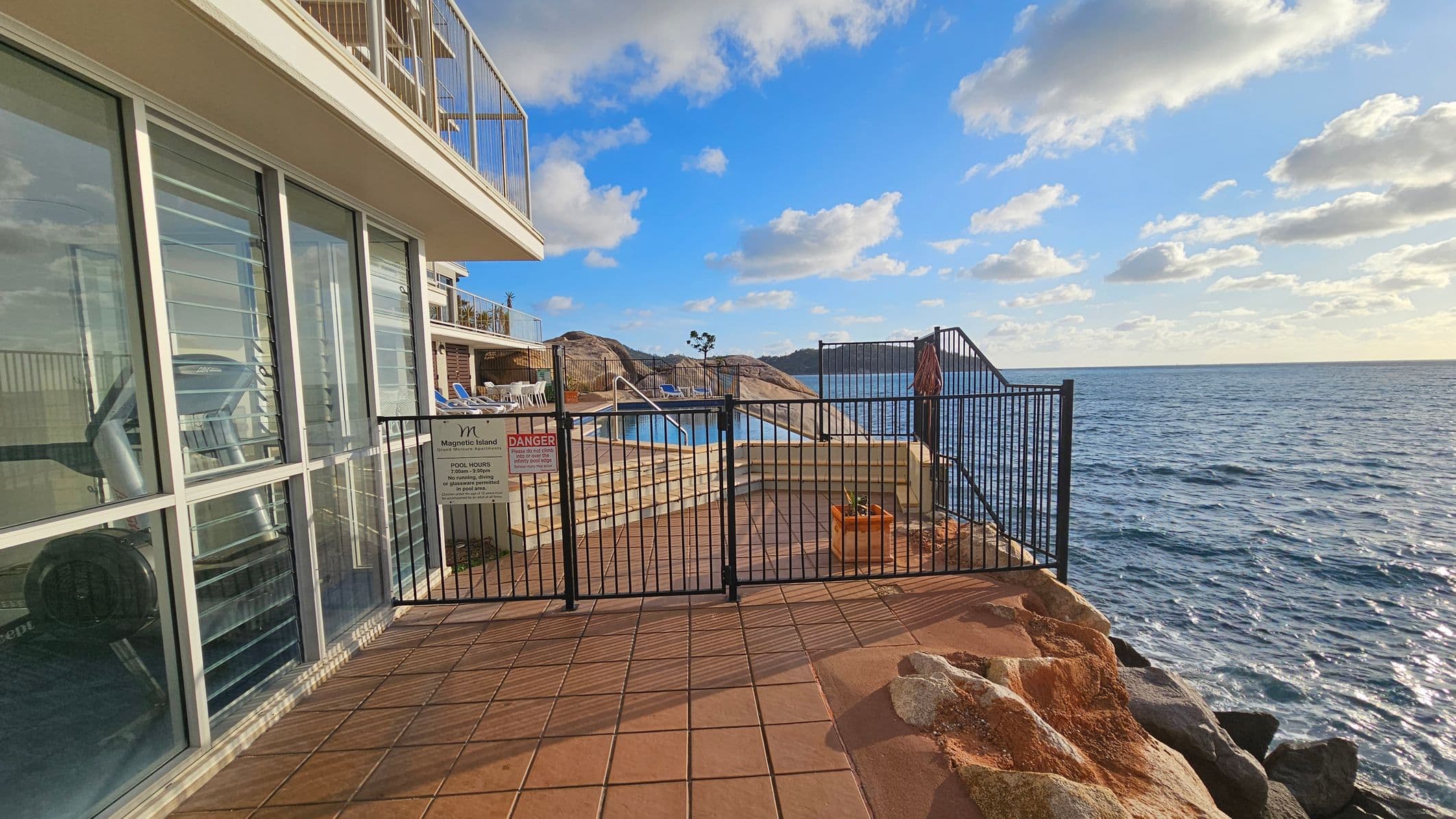 A coastal view with a building on the left featuring large glass windows. In the centre, a black metal gate with signs leads to a pool area with steps and a few sun loungers. The background shows rocky terrain and the ocean extending to the right. The foreground has a tiled floor and some rocks.
