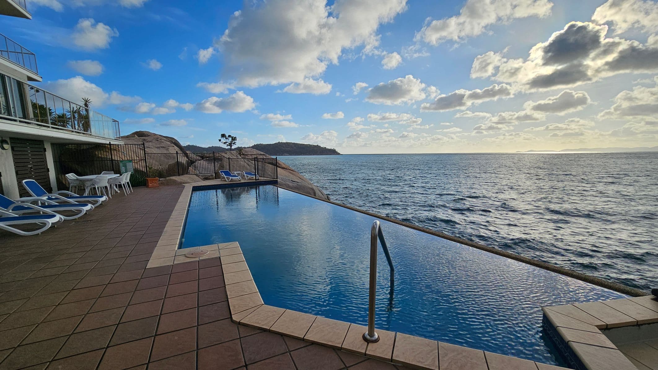 An infinity pool with a metal handrail is in the foreground, overlooking a vast ocean. On the left, there are several white sun loungers and a table with chairs on a tiled patio. A building with glass railings is adjacent to the patio. In the background, rocky terrain and a distant landmass are visible. The sky is partly cloudy.