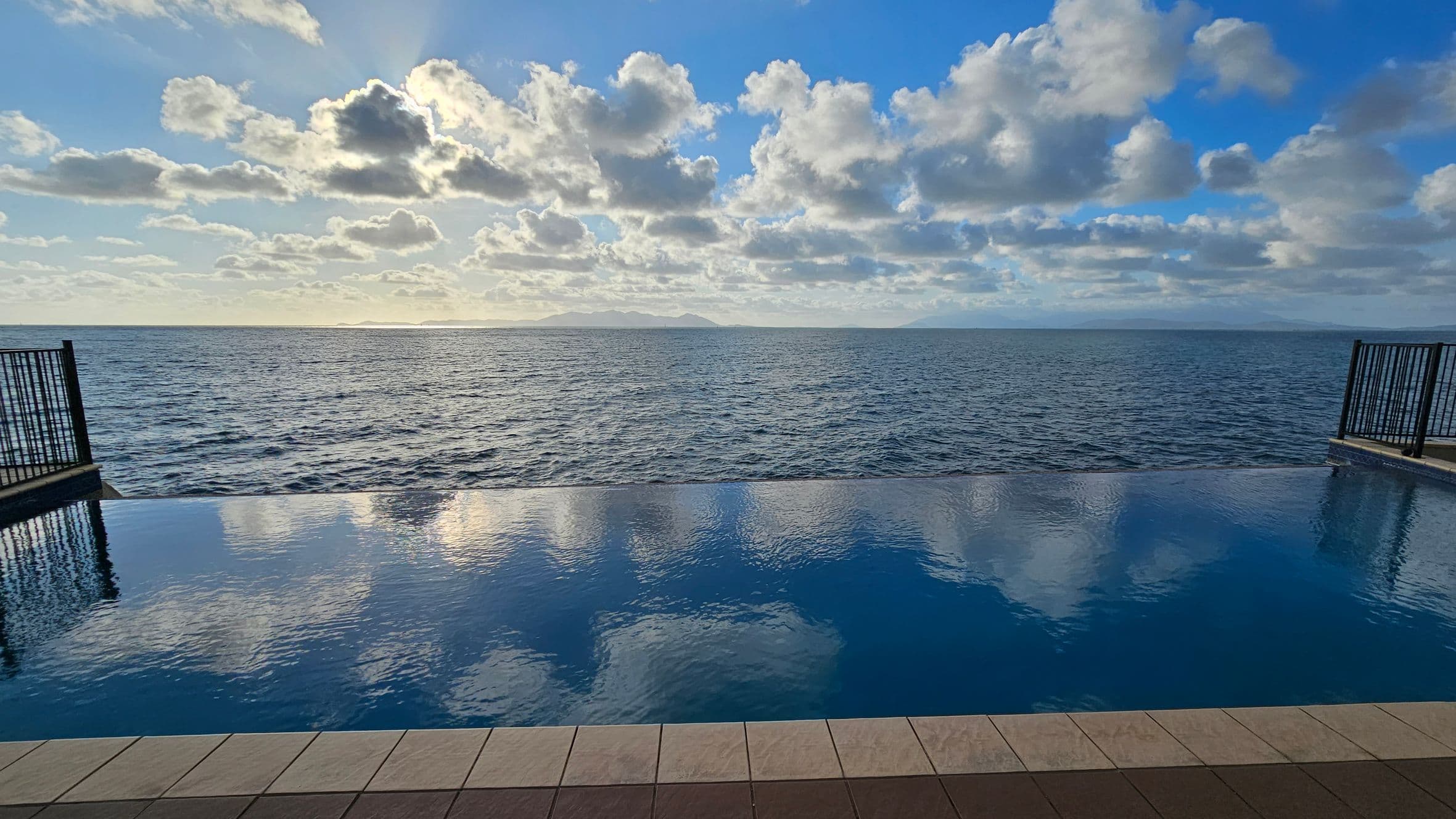 An infinity pool in the foreground reflects the sky and clouds. The pool is bordered by a tiled deck with a pattern of light and dark tiles. On the left and right, black metal railings frame the view. In the background, the ocean stretches to the horizon, meeting a sky filled with scattered clouds. Distant landforms are faintly visible on the horizon.