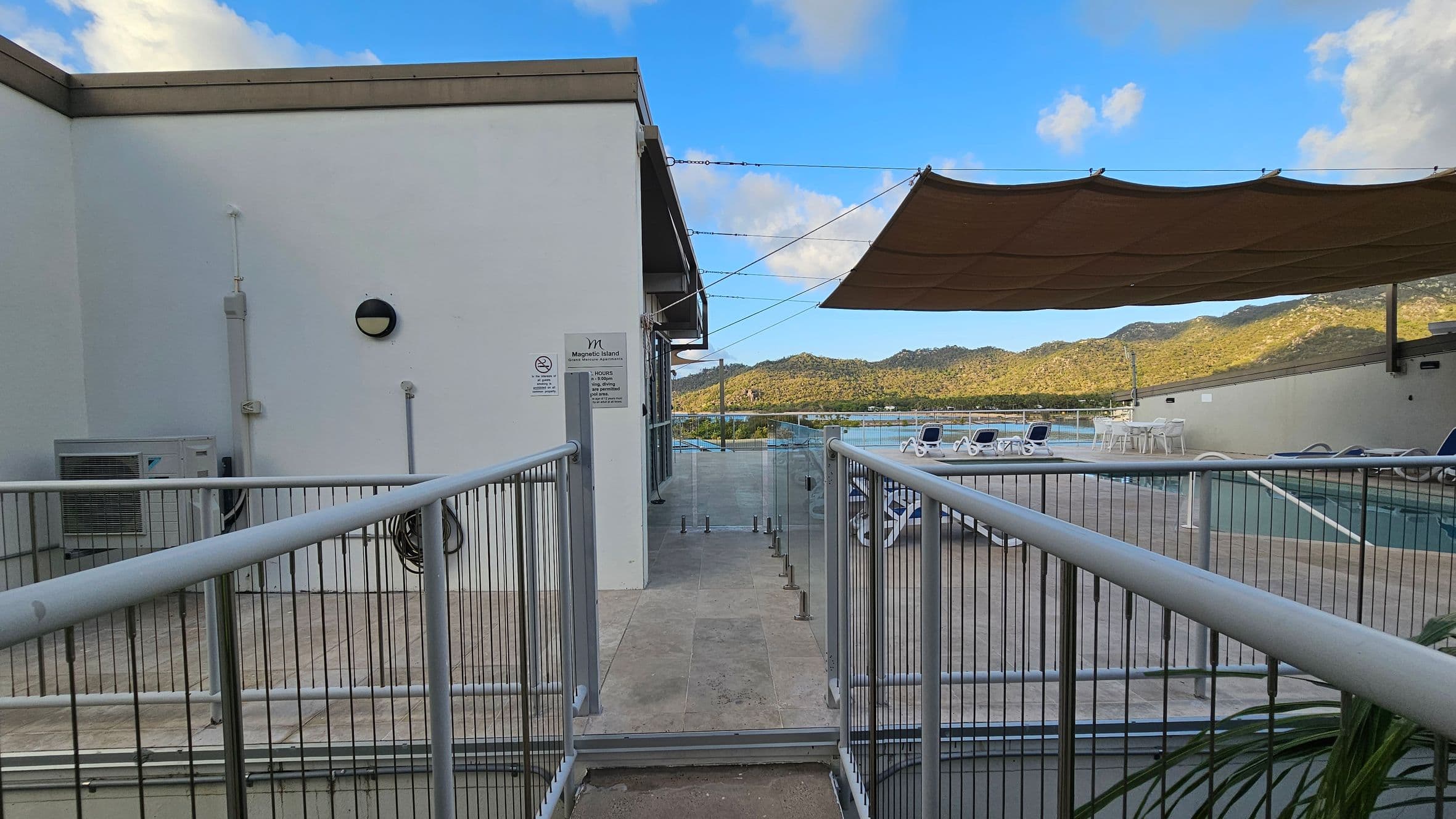 A rooftop area with a fenced walkway leading to a pool deck. On the left, a white wall with a round light fixture and an air conditioning unit. The right side features a shaded area with a large canopy, several sun loungers, and a table with chairs. In the background, there are green hills and a glimpse of water. The floor is tiled, and glass barriers line the pool area.