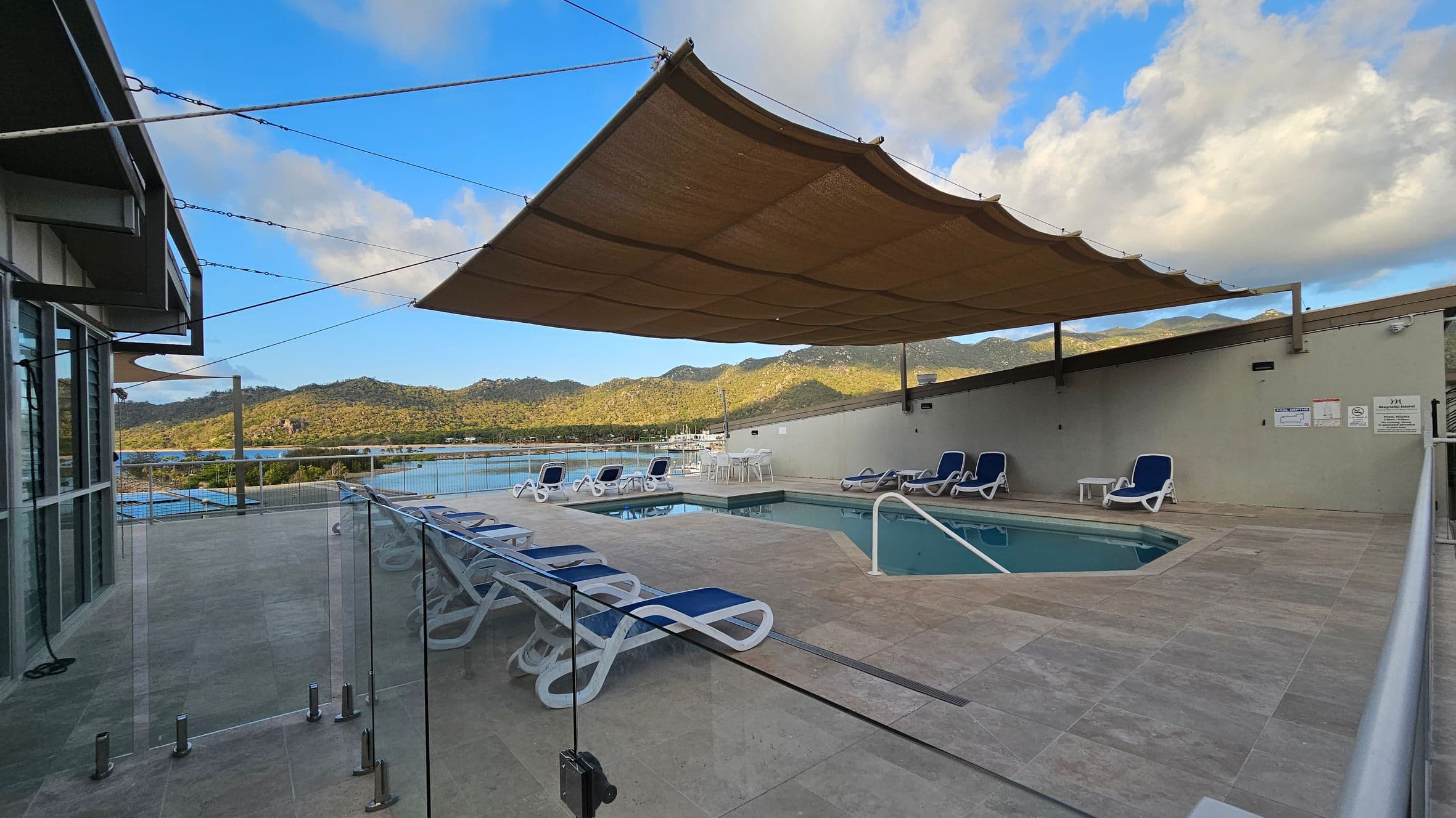 A rooftop pool area with a rectangular pool in the centre, surrounded by blue and white sun loungers. A large beige shade sail is suspended above. Glass barriers line the left side, with a view of distant hills and water. The floor is tiled in light grey, and there are small tables between some loungers. A green wall with signs is on the right.