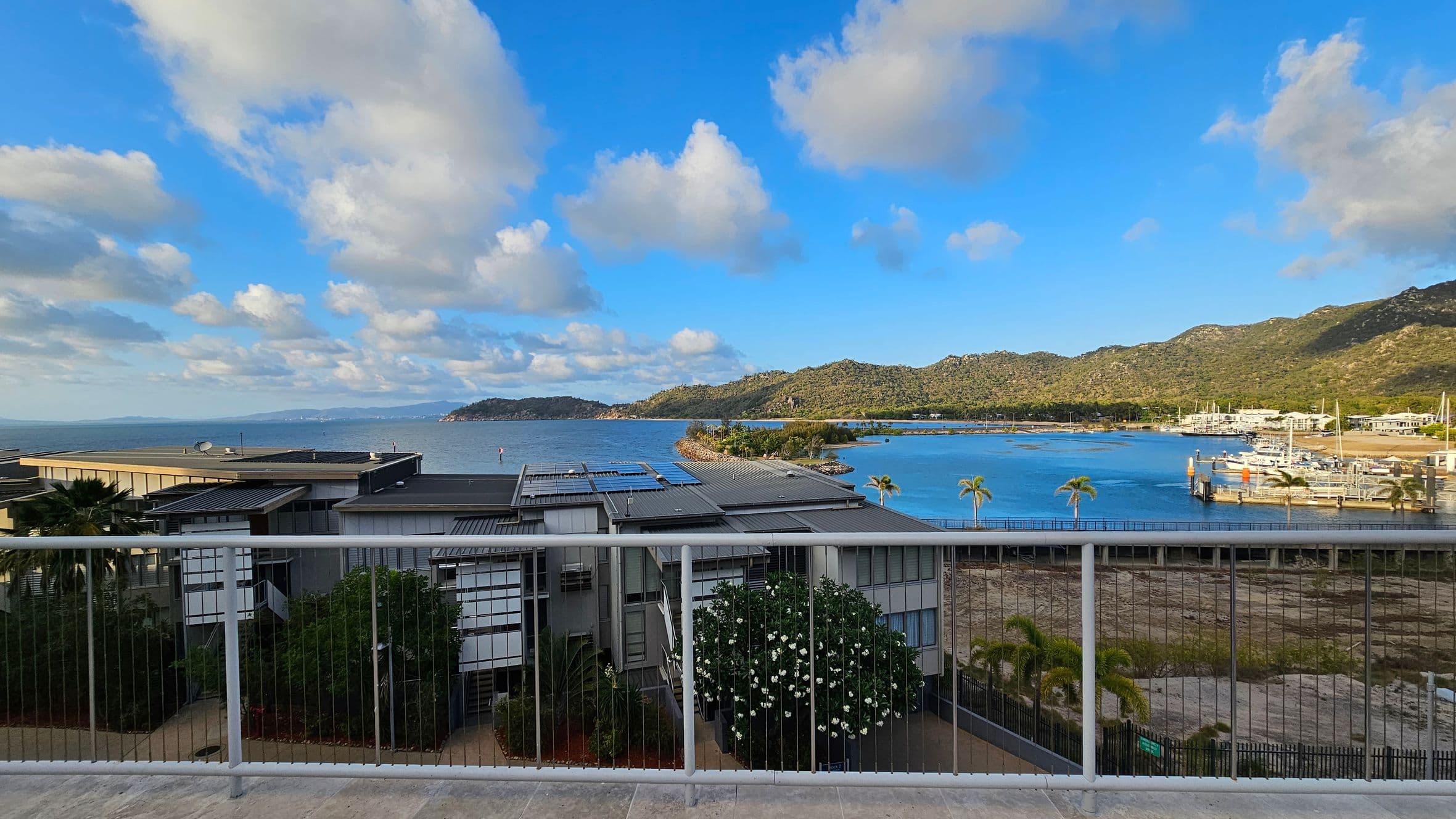 A view from a balcony with a metal railing in the foreground. Below, modern buildings with flat roofs and solar panels are visible. In the background, a body of water stretches out, bordered by a mountainous landscape. To the right, a marina with several boats is docked. Palm trees are scattered throughout the scene, adding a tropical feel. The sky is mostly clear with a few scattered clouds.