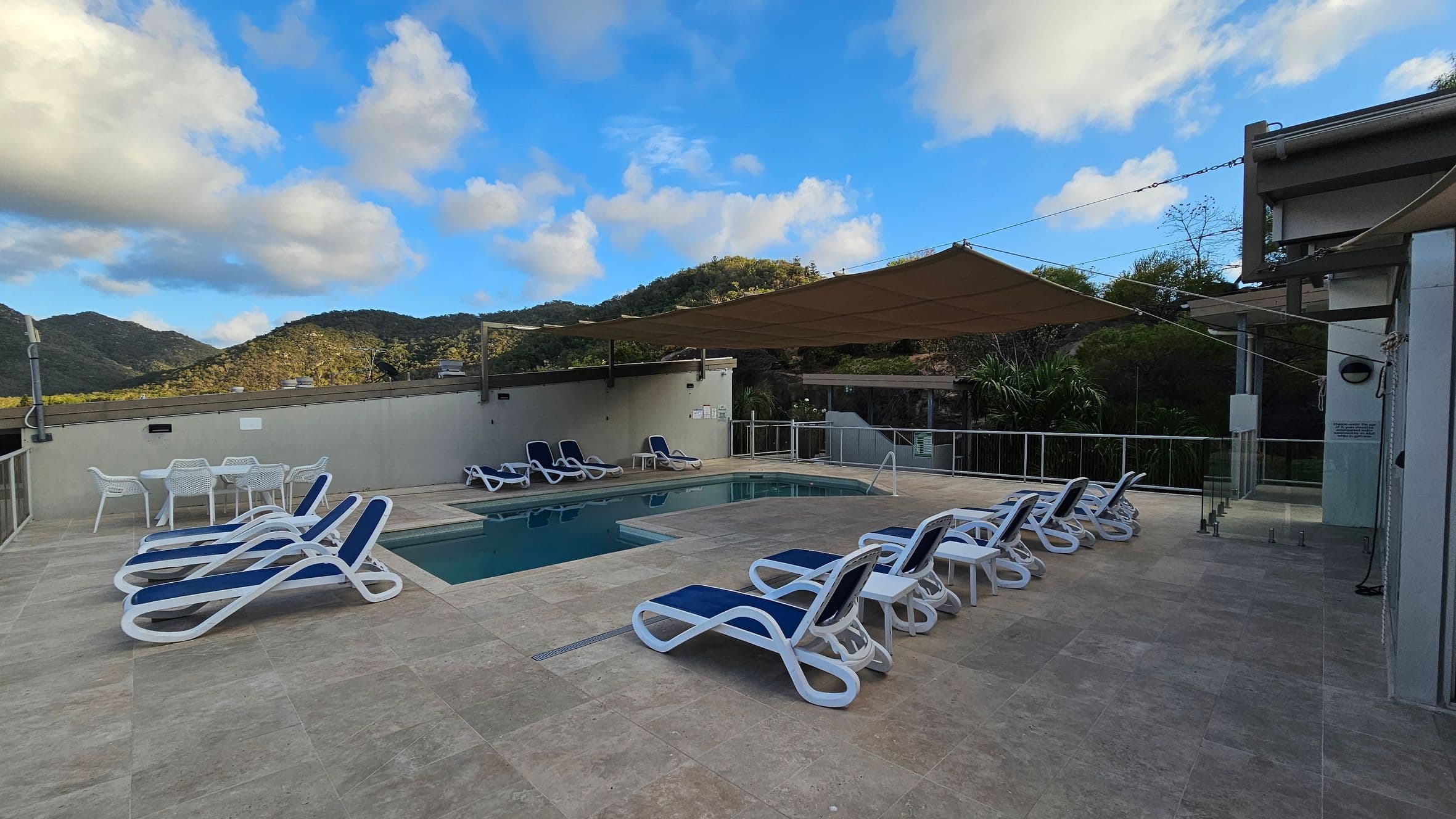 A pool area with several white and blue sun loungers arranged around a rectangular swimming pool. On the left, there is a white table with chairs. The ground is paved with light grey tiles. In the background, there are green hills and a partly cloudy sky. A beige shade sail is stretched over part of the pool area, and a glass railing encloses the space.