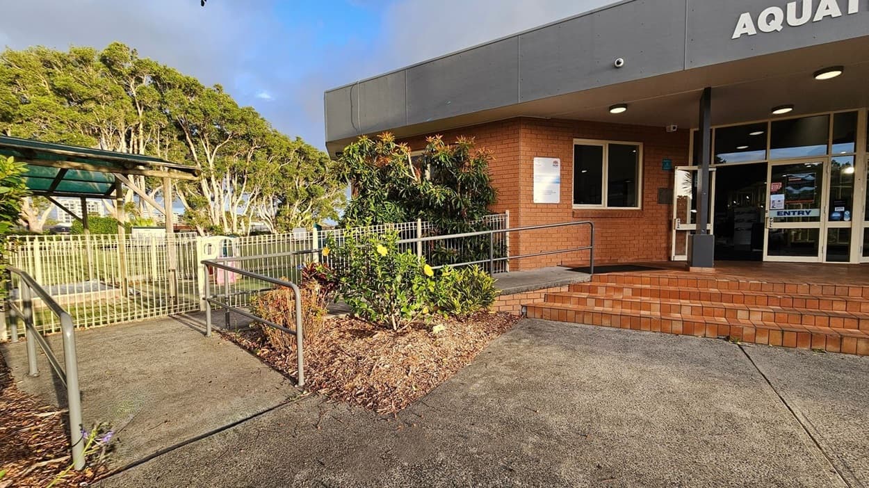A building entrance with a sign reading "AQUAT" is on the right, featuring glass doors and brick steps. A ramp with metal railings leads up to the entrance. In the centre, there are bushes with yellow flowers. On the left, a fenced area with a green structure and trees is visible. The ground is paved with concrete, and the building has a grey overhang with lights.