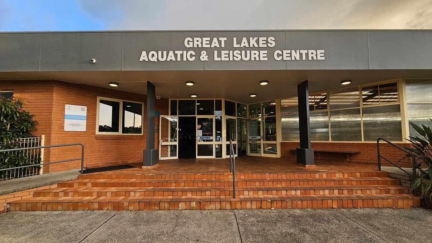 Entrance of the Great Lakes Aquatic & Leisure Centre with a grey overhang displaying the centre's name in white letters. The building is made of red brick with large glass doors and windows. A set of brick steps leads up to the entrance, flanked by metal railings. A sign is visible on the left wall, and there are plants on either side of the entrance. The pavement in front is concrete.