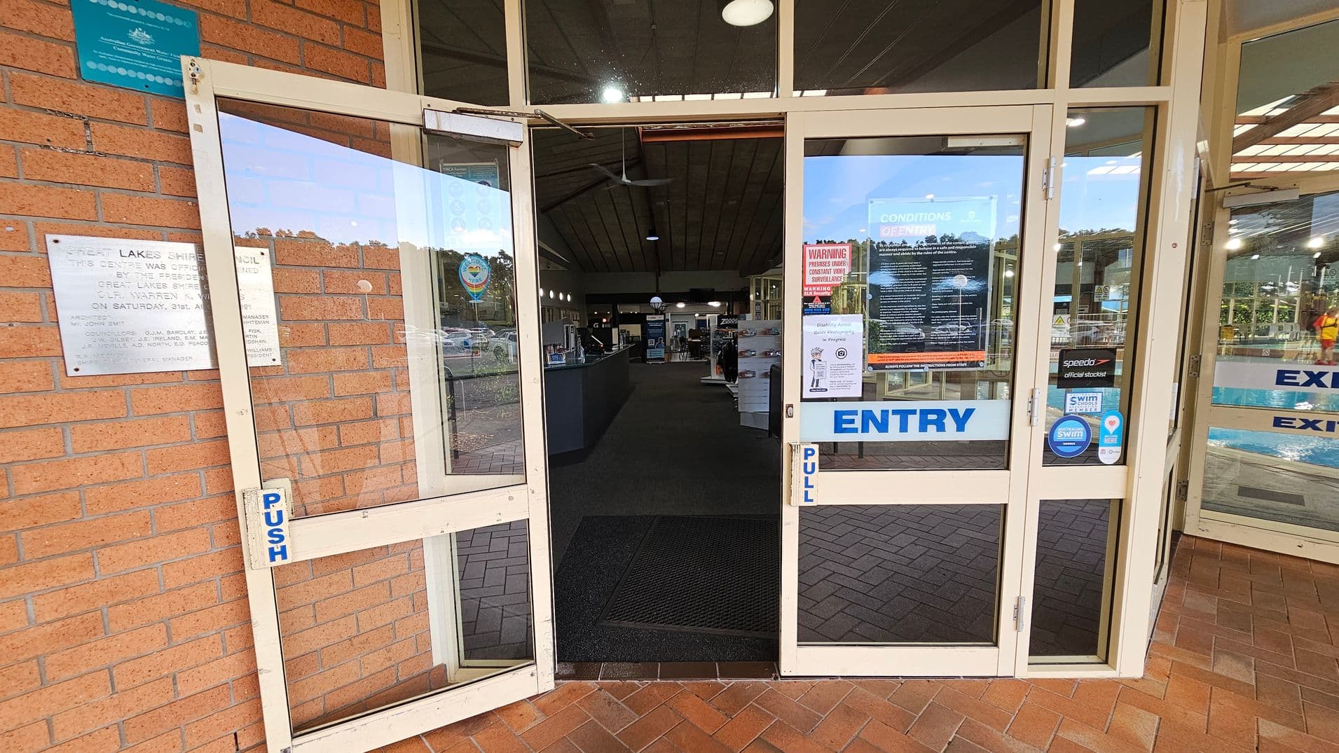 Glass entrance doors to a building with "ENTRY" and "EXIT" signs. The left door is open, showing a brick wall with a plaque. The right door is also open, displaying various notices and stickers. Inside, a reception area with a counter and informational displays is visible. The floor is tiled with a herringbone pattern, and the ceiling has a series of lights.