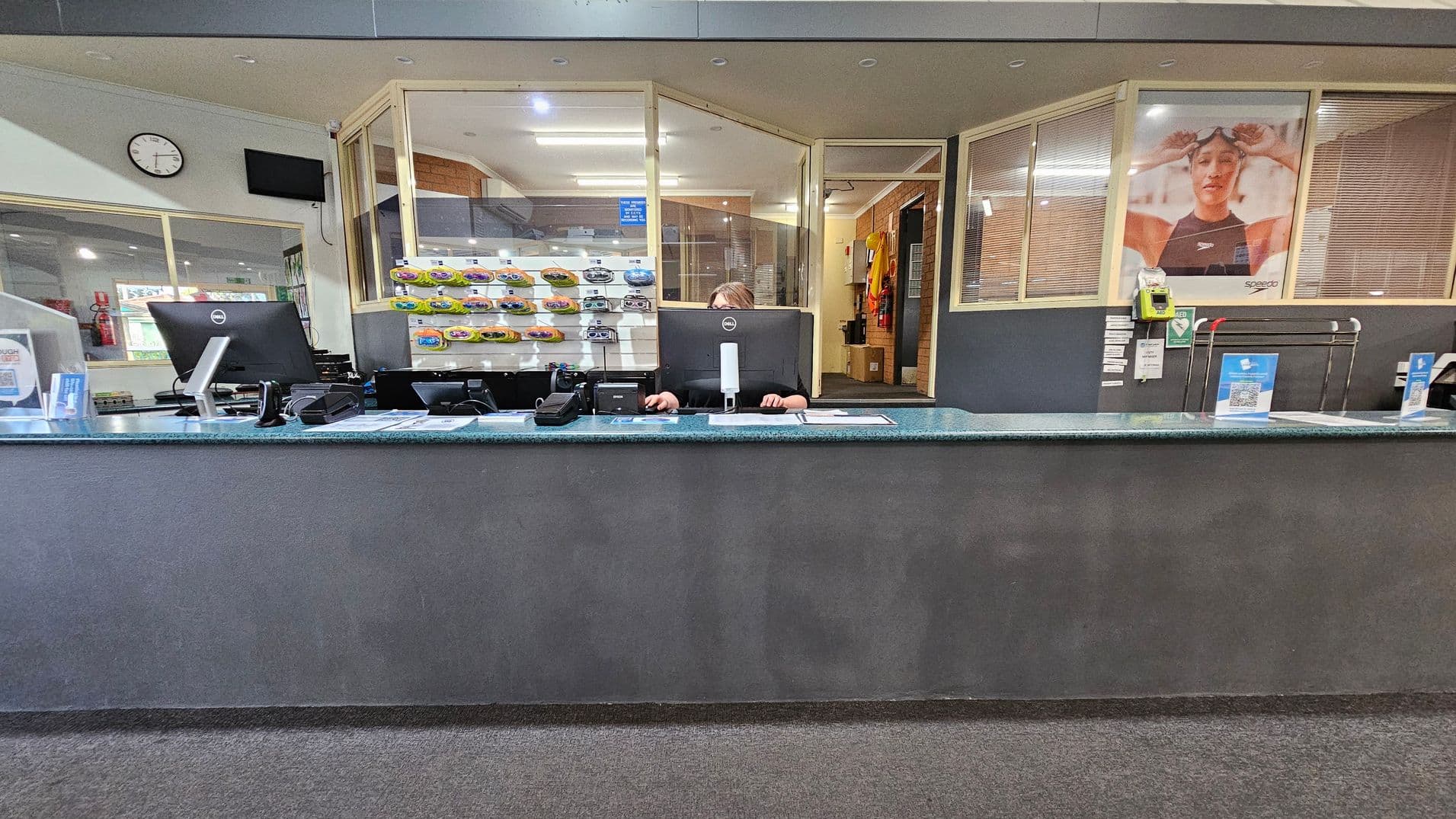 A reception desk with a grey front and a green countertop. Two computer monitors are on the desk, with a person seated behind them. On the left, a wall clock and a small TV screen are mounted. Behind the desk, shelves display various items. On the right, a large poster of a swimmer wearing goggles is visible. The floor is carpeted, and the ceiling has recessed lighting.