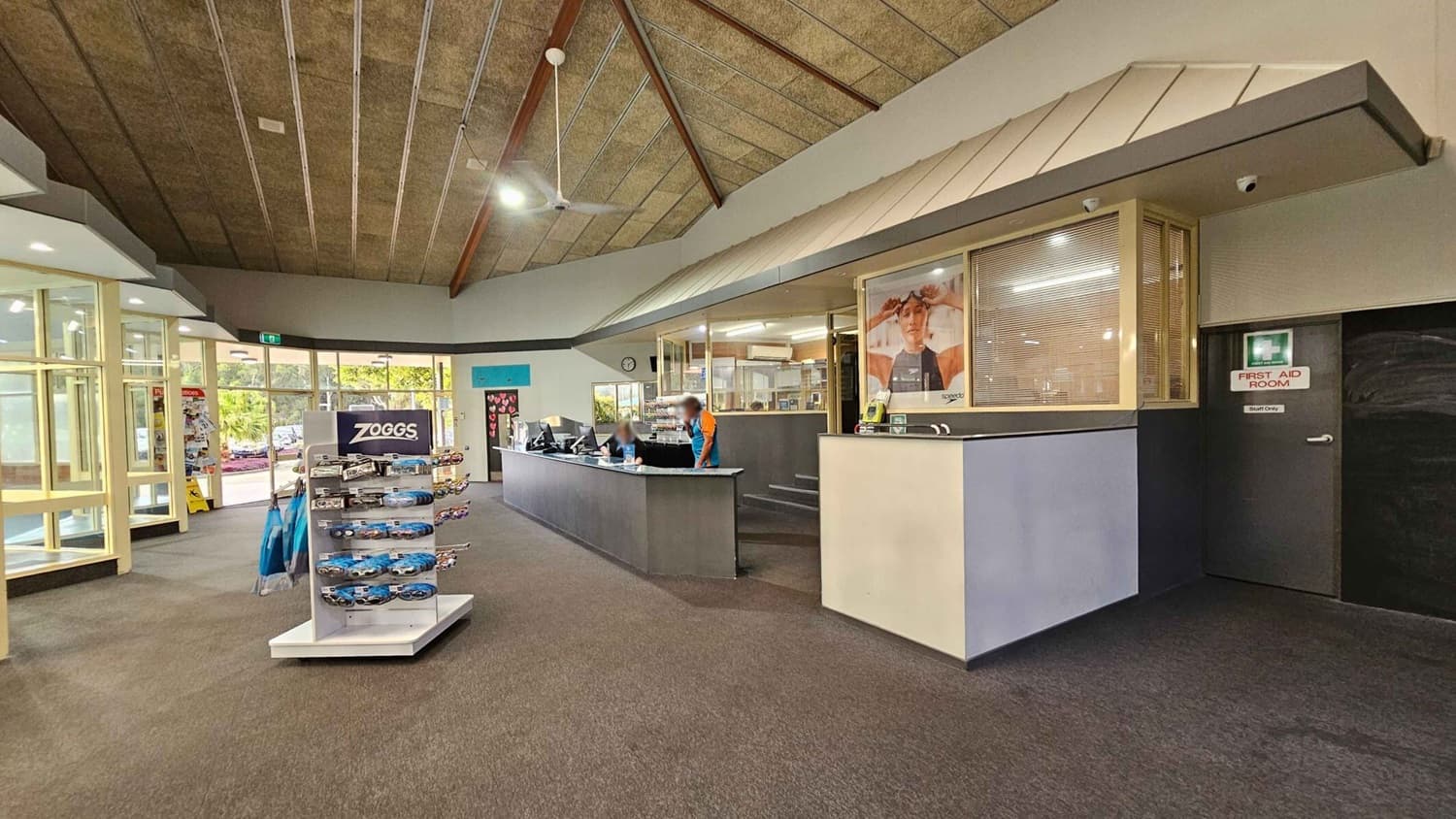 Reception area with a high, angled ceiling featuring exposed beams. On the left, a display stand with swimming goggles and accessories. In the centre, a long reception desk with two staff members working. Behind the desk, a large window and a poster. To the right, a door marked "First Aid Room." The floor is carpeted, and the entrance is visible in the background with glass doors and a view outside.