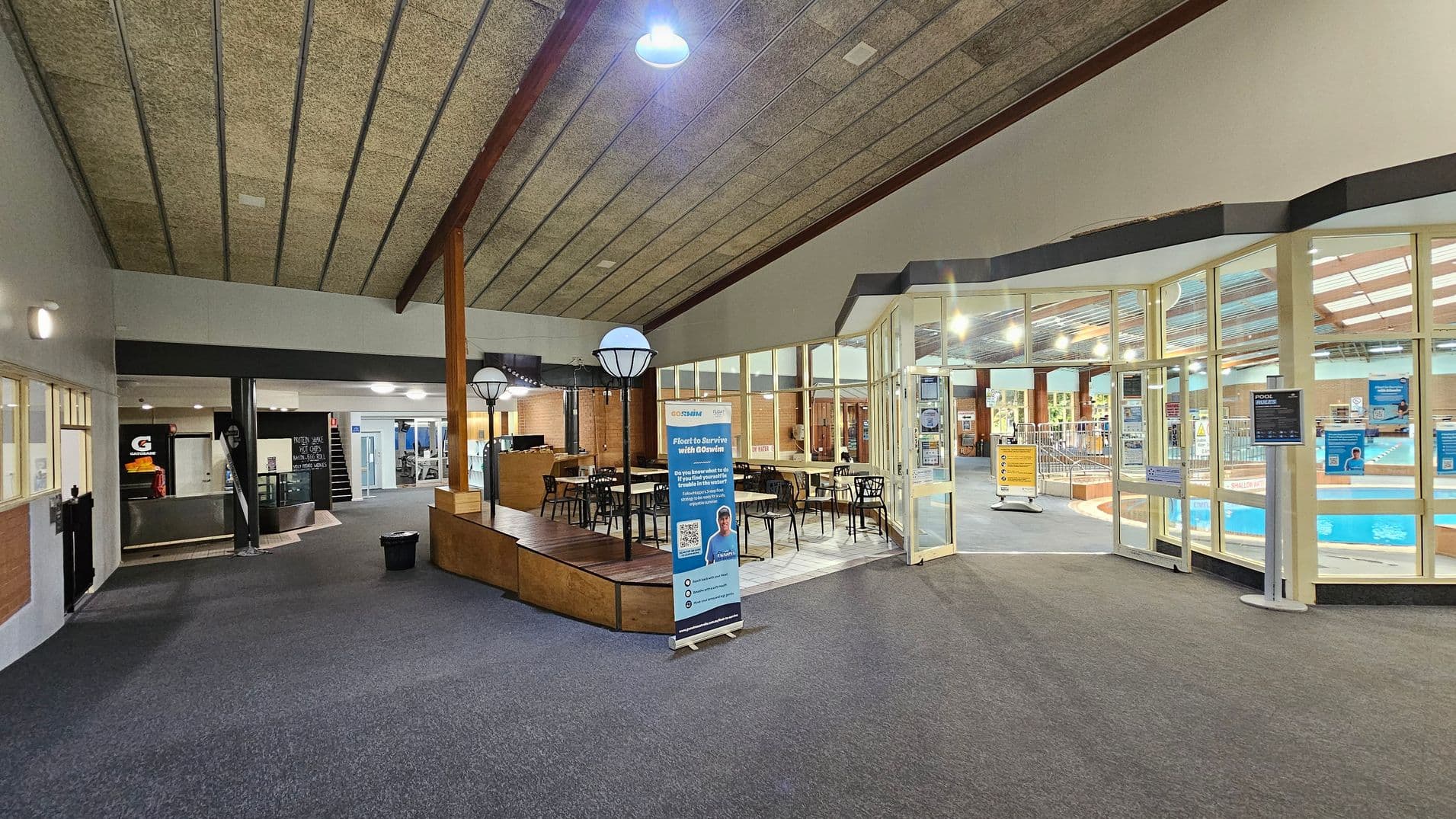 A spacious indoor area with a high, textured ceiling. On the left, there's a reception area with a counter and a blackboard displaying information. In the centre, a seating area with tables and chairs is visible, along with a standing banner. On the right, large glass windows and doors reveal a swimming pool area with bright lighting. The floor is carpeted, and there are wooden accents throughout the space.