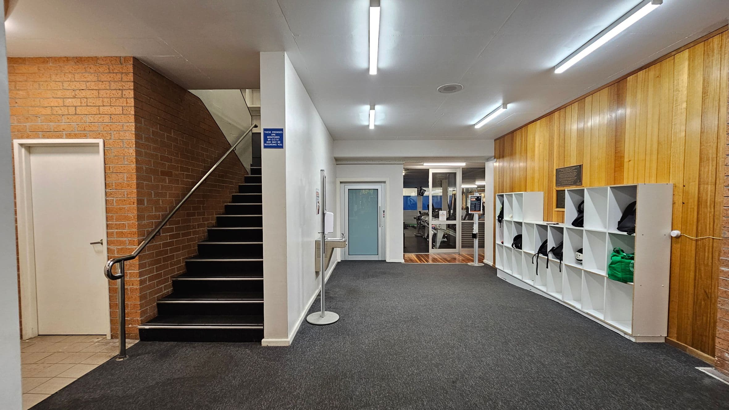 A hallway with a dark carpeted floor and fluorescent ceiling lights. On the left, there's a staircase with a metal handrail and a closed door with a tiled floor area. In the centre, a blue door is visible, leading to a room with gym equipment. On the right, a wooden-panelled wall features a set of white cubby shelves containing bags and a green hoodie.