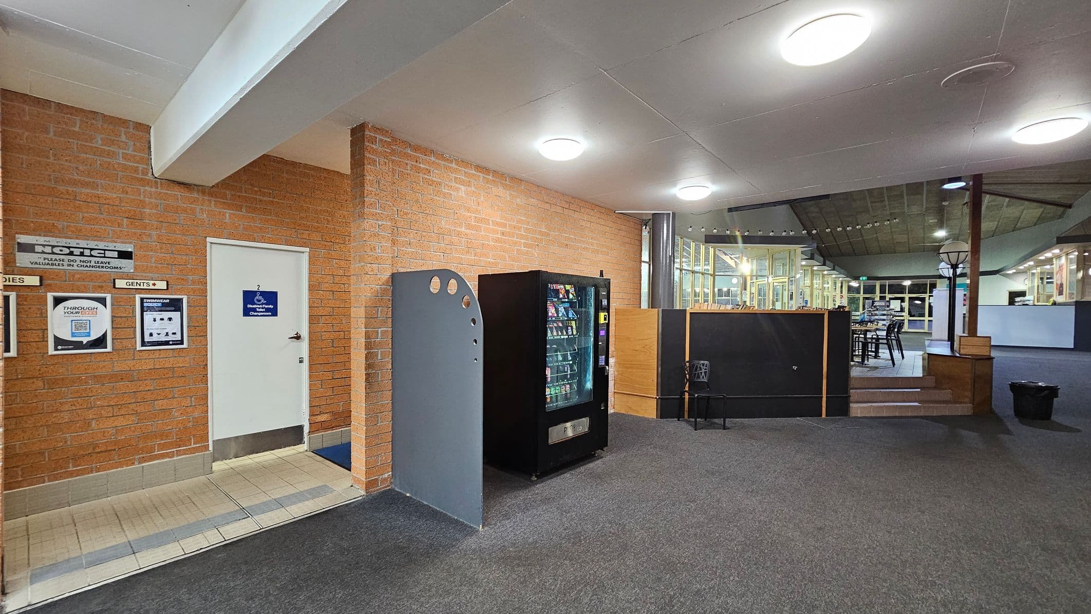 A room with a grey carpeted floor and a white ceiling with circular lights. On the left, there's a brick wall with a white door labelled "Gents" and "Disabled Change Room". Notices and signs are mounted on the wall. A vending machine is positioned next to a curved partition. On the right, a raised area with tables and chairs is visible, separated by a low wall. A coat rack stands nearby, and large windows are in the background.