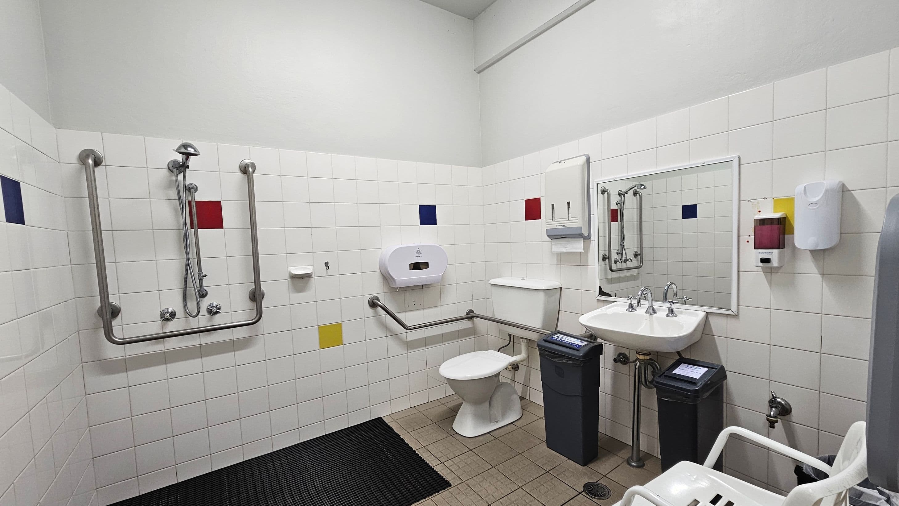 A bathroom with white tiled walls featuring red, blue, and yellow accent tiles. On the left, there is a shower area with grab bars and a handheld showerhead. In the centre, a toilet with support rails and a toilet paper dispenser is visible. On the right, a sink with dual taps is mounted below a mirror, with soap and paper towel dispensers nearby. The floor is tiled with a black mat near the shower area. A white chair is partially visible in the foreground.