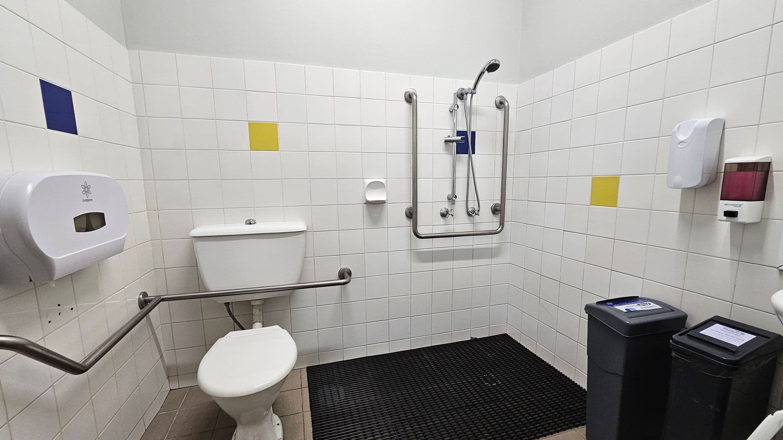 A public bathroom with white tiled walls featuring occasional blue and yellow tiles. On the left, there's a toilet with a metal grab bar and a toilet paper dispenser. In the centre, a shower area with a handheld showerhead and additional grab bars. On the right, a soap dispenser and a hand sanitiser are mounted on the wall. Two black bins with lids are on the floor, which is covered with a black rubber mat. The ceiling is plain white.