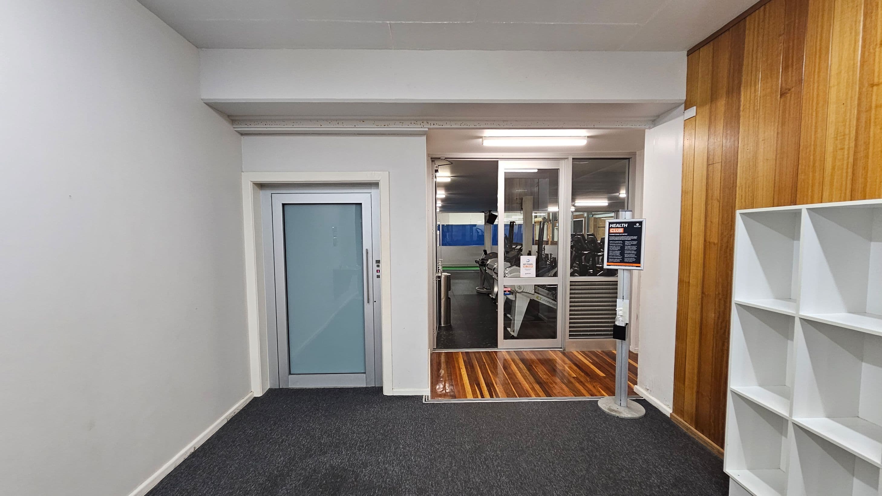 A hallway with a dark carpeted floor and fluorescent ceiling lights. On the left, a staircase with a metal handrail ascends beside a brick wall. A white door is adjacent to the stairs. In the centre, a blue door is visible, leading to a room with gym equipment. On the right, a wooden panelled wall features a plaque above white cubby shelves holding bags and a green item.