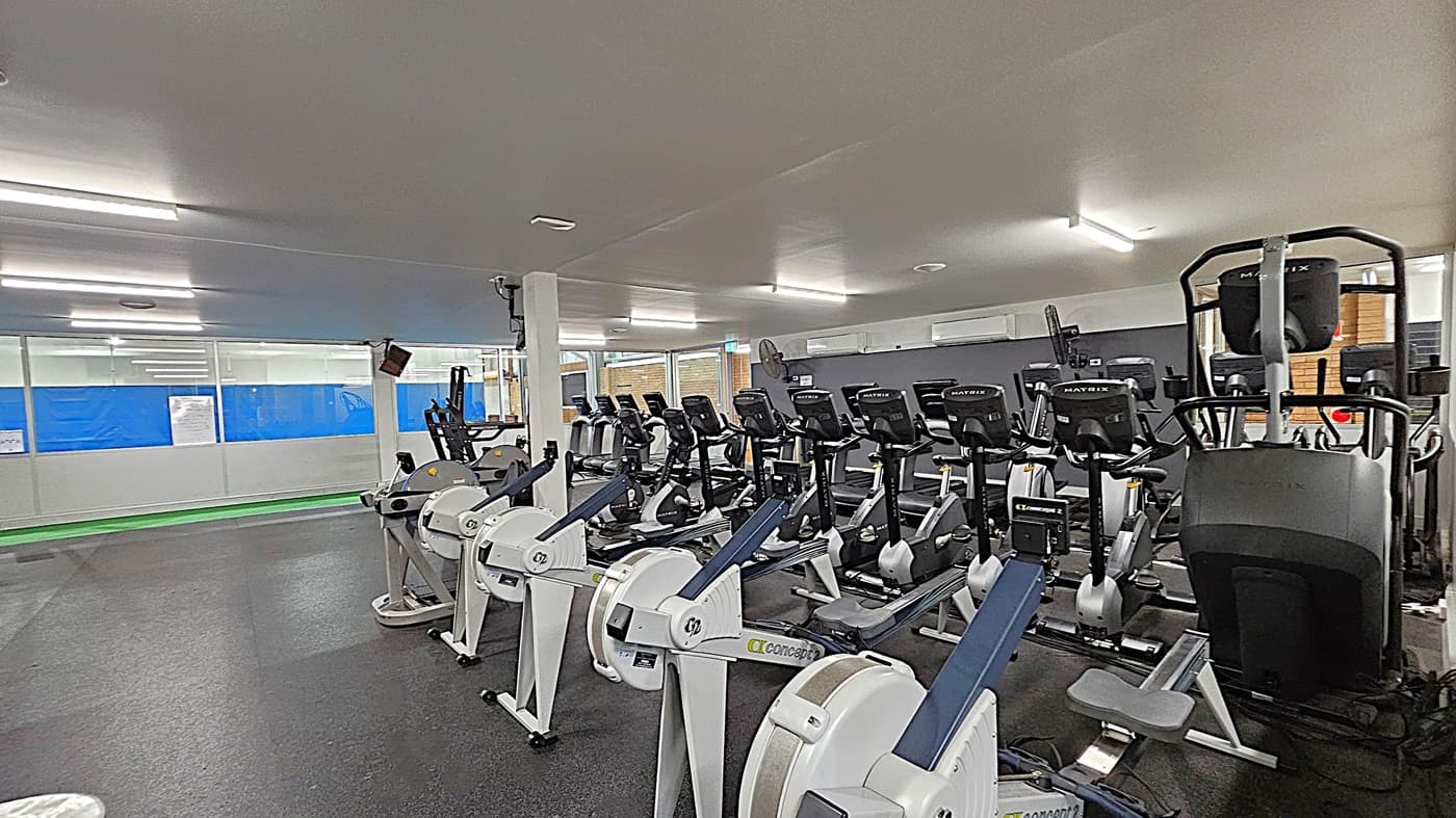 A gym with a row of rowing machines in the foreground, each equipped with a digital display. Behind them, there are several stationary bikes lined up. The floor is covered with dark rubber matting, and the ceiling is fitted with fluorescent lights. On the left, large windows with blue coverings allow some natural light. A white pillar stands near the centre, and there are air conditioning units on the back wall.