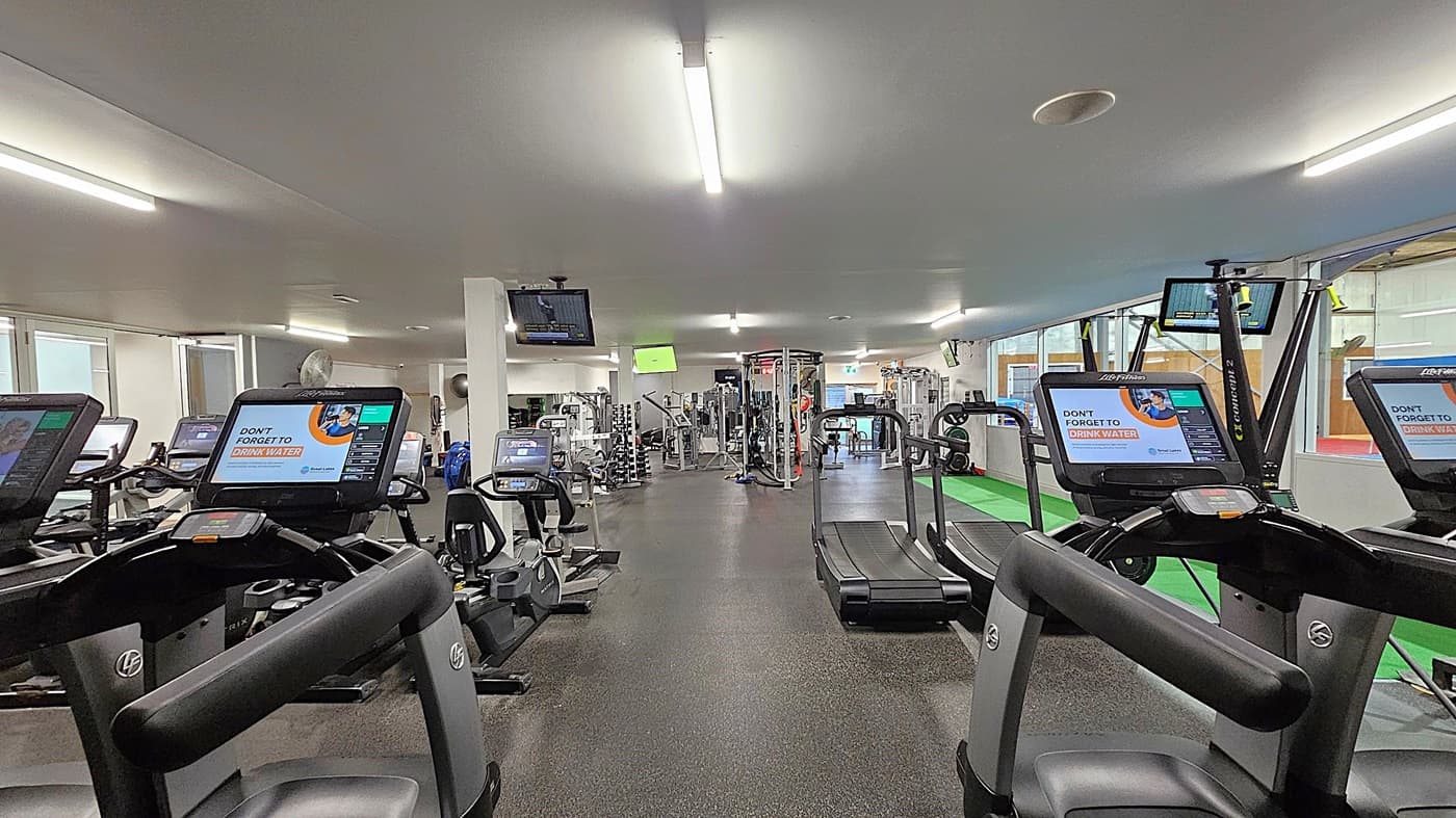 A gym interior with exercise machines. In the foreground, several treadmills and exercise bikes have screens displaying a reminder to drink water. The floor is dark and rubberised, while the ceiling is white with fluorescent lights. In the background, there are various weight machines and equipment. Large windows line the right side, and a green mat is visible near the machines. Several wall-mounted TVs are also present.