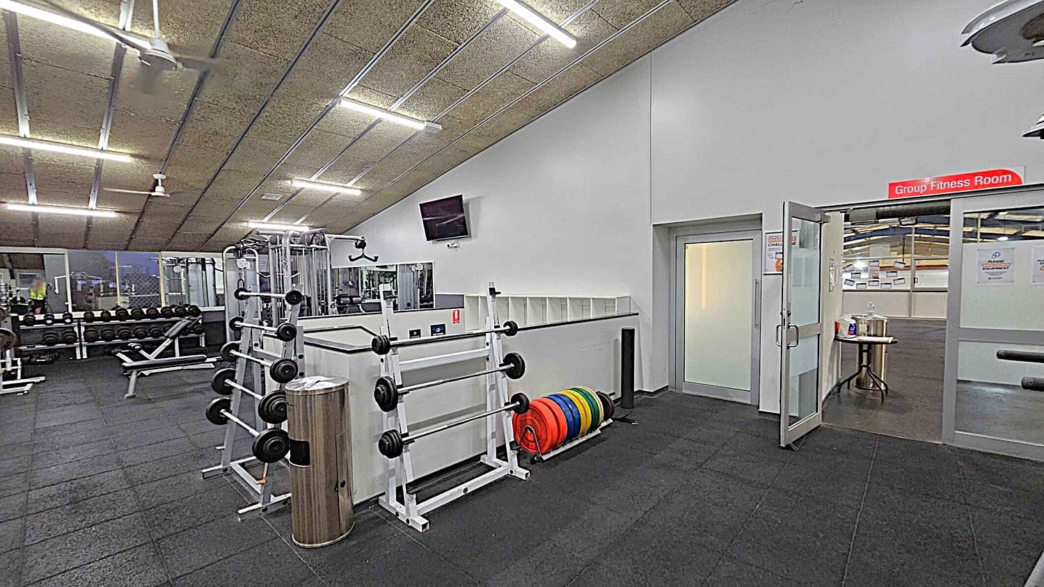 A gym interior with a sloped ceiling featuring fluorescent lights and a ceiling fan. On the left, there are weight racks with dumbbells and benches. In the centre, a rack holds barbells and weight plates. On the right, a door marked "Group Fitness Room" is open, revealing a table with cleaning supplies. The floor is covered with black rubber tiles.