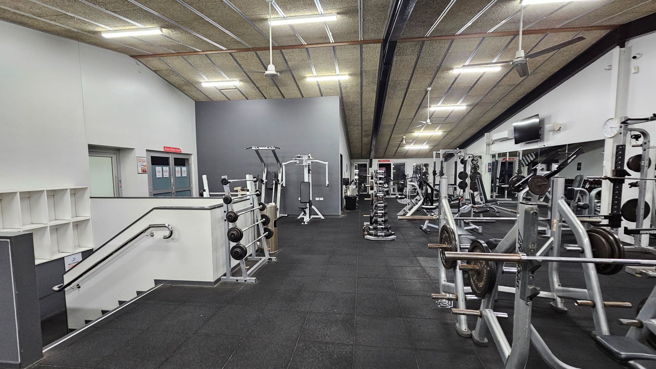 A gym interior with various exercise equipment. On the left, there are stairs with a handrail leading down, and a wall with cubby shelves. In the centre, there are weight machines and a row of dumbbells. On the right, there are more weightlifting machines and benches. The ceiling has exposed beams and fluorescent lights, and the floor is covered with black rubber mats.