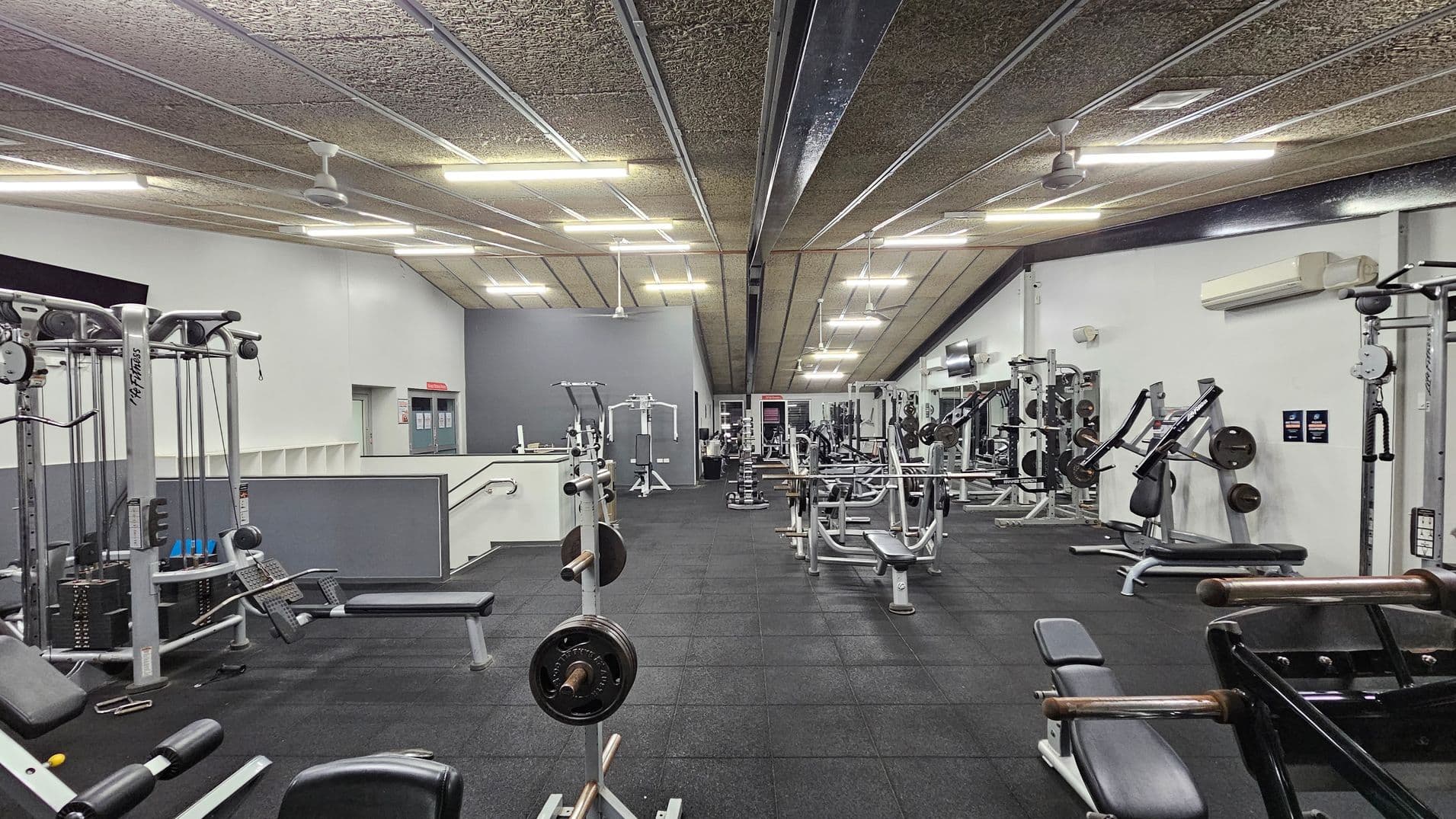 A spacious gym with various exercise equipment, including weight machines and benches, arranged neatly on a black rubber floor. The ceiling is lined with fluorescent lights and ceiling fans. On the left, there's a staircase leading to a raised area. The walls are painted white and grey, with air conditioning units visible on the right. The gym is well-lit and organised, with ample space between the equipment.