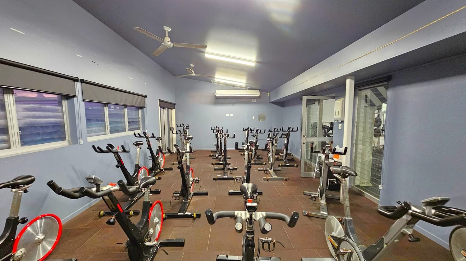 A room with multiple stationary exercise bikes arranged in rows on a brown tiled floor. The left wall has several windows with grey blinds. The ceiling is grey with fluorescent lights and ceiling fans. On the right, there's a glass door leading to another area. An air conditioning unit is mounted on the back wall.