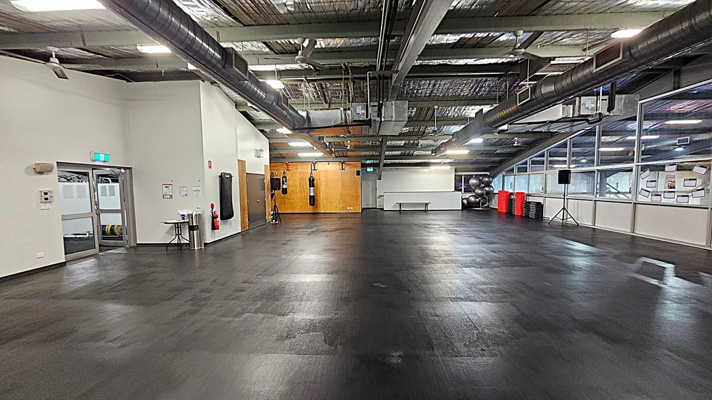 A spacious gym with a dark, polished floor and an industrial ceiling with exposed ducts. On the left, there are doors and a small table with a fire extinguisher nearby. In the centre, a wooden wall with hanging punching bags. On the right, large windows with exercise balls and stacked red and black step platforms. A speaker on a stand is also visible. The room is well-lit with fluorescent lights.
