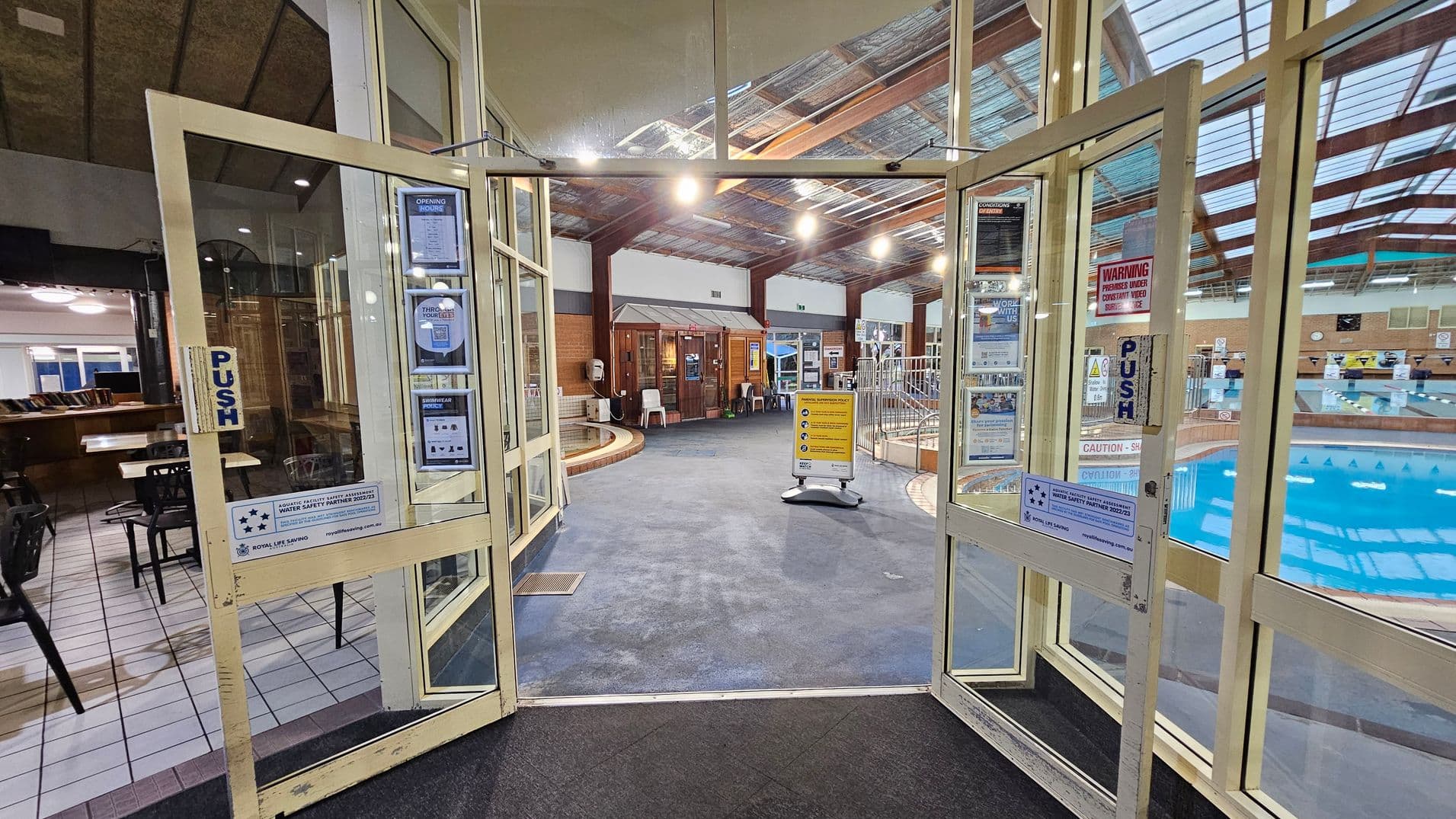 Glass doors with "PUSH" signs open into an indoor swimming pool area. On the left, there is a seating area with tables and chairs on a tiled floor. In the centre, a carpeted walkway leads to a wooden structure with chairs and posters. On the right, a pool with blue water is visible, surrounded by a railing. The ceiling is made of transparent panels supported by wooden beams, allowing natural light to illuminate the space.