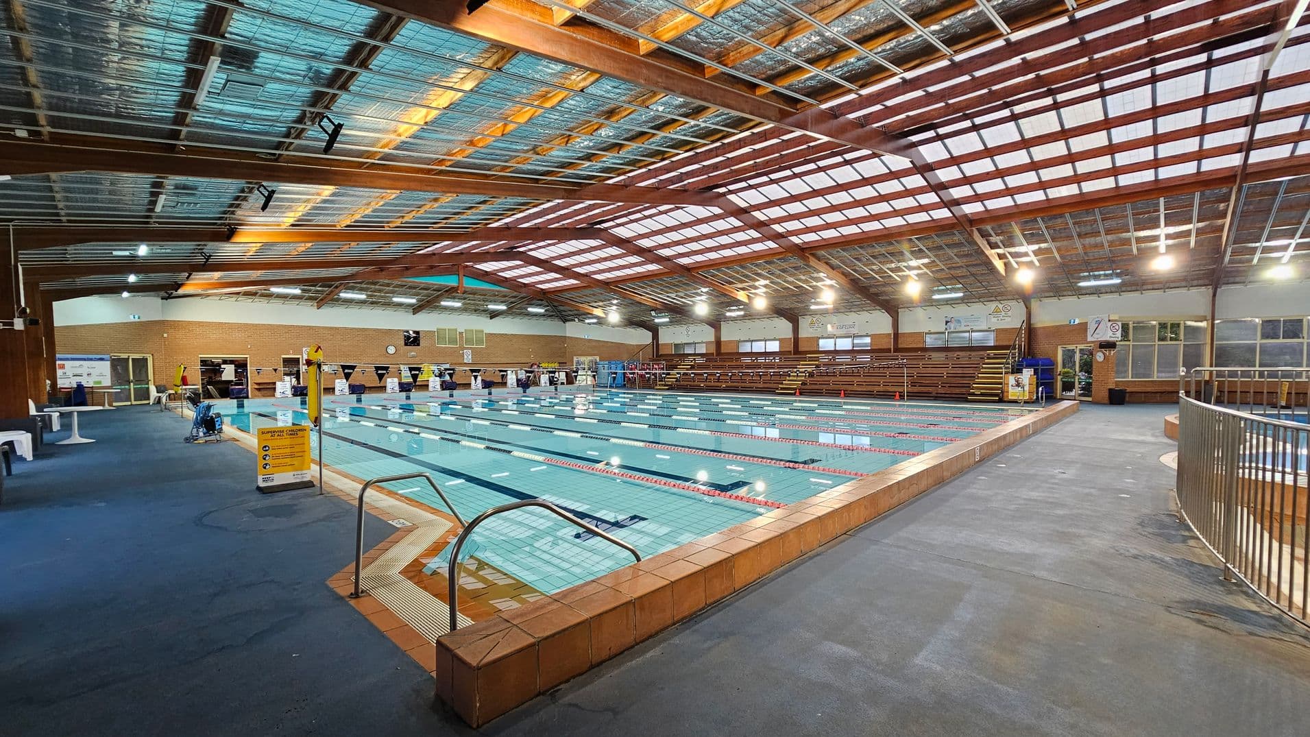An indoor swimming pool with multiple lanes, surrounded by a tiled deck. On the left, there are white benches and tables on a blue carpeted floor. The right side features a metal railing and a grey floor. The background shows brick walls with windows and a clock. The ceiling is made of wooden beams and translucent panels, allowing natural light to filter through. Overhead lights are evenly spaced across the ceiling.