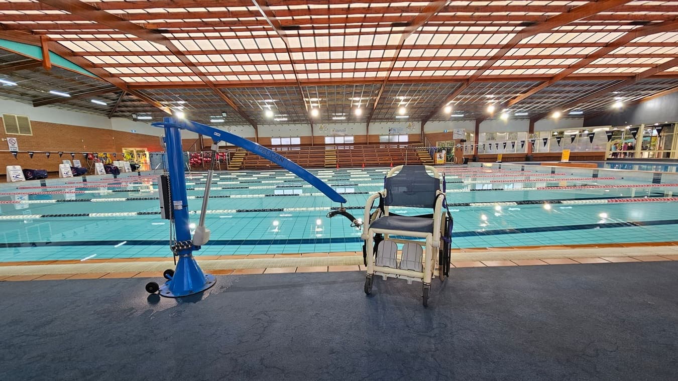 A swimming pool with multiple lanes is in the centre, surrounded by a tiled deck. On the left, a blue pool lift is installed, and next to it on the right is a wheelchair. The background features wooden bleachers and large windows. The ceiling is made of wooden beams with a grid of translucent panels, allowing light to filter through. The floor near the pool is covered with a dark mat.