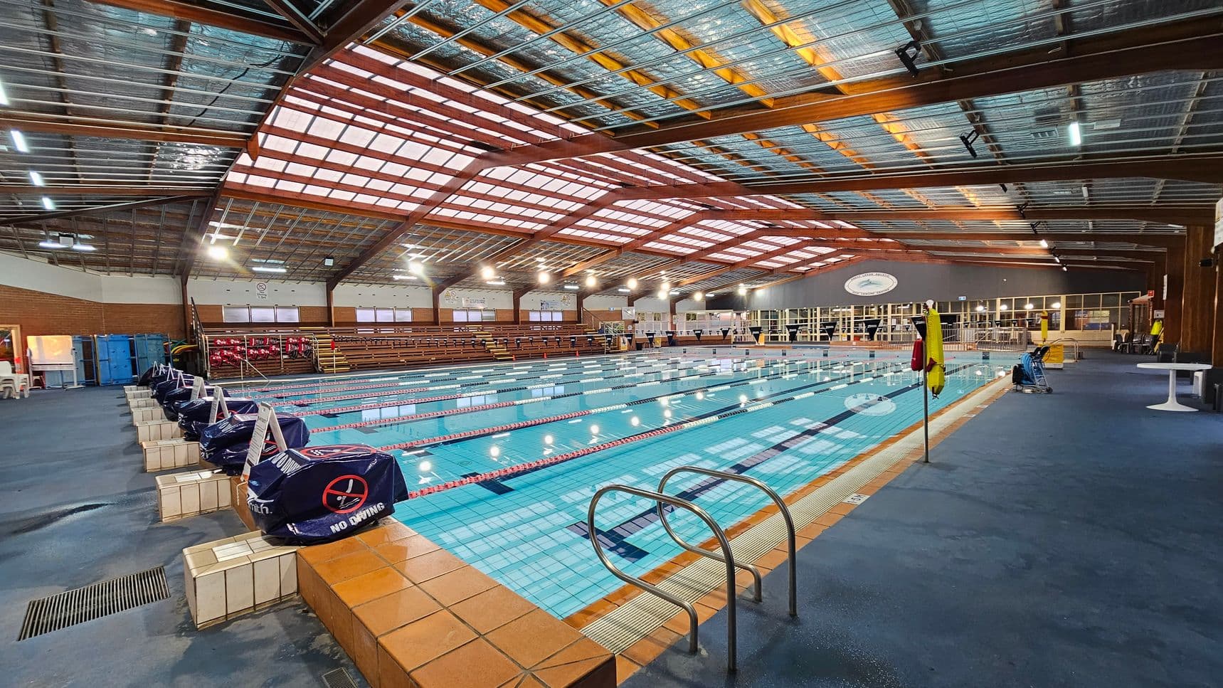 An indoor swimming pool with multiple lanes, surrounded by a blue-tiled deck. On the left, there are covered starting blocks and a row of wooden bleachers. The ceiling is made of metal and wood beams with skylights. On the right, there are tables and chairs near large windows. The pool has a metal ladder on the right side, and the floor is covered with blue carpet. The lighting is bright, with numerous overhead lights.