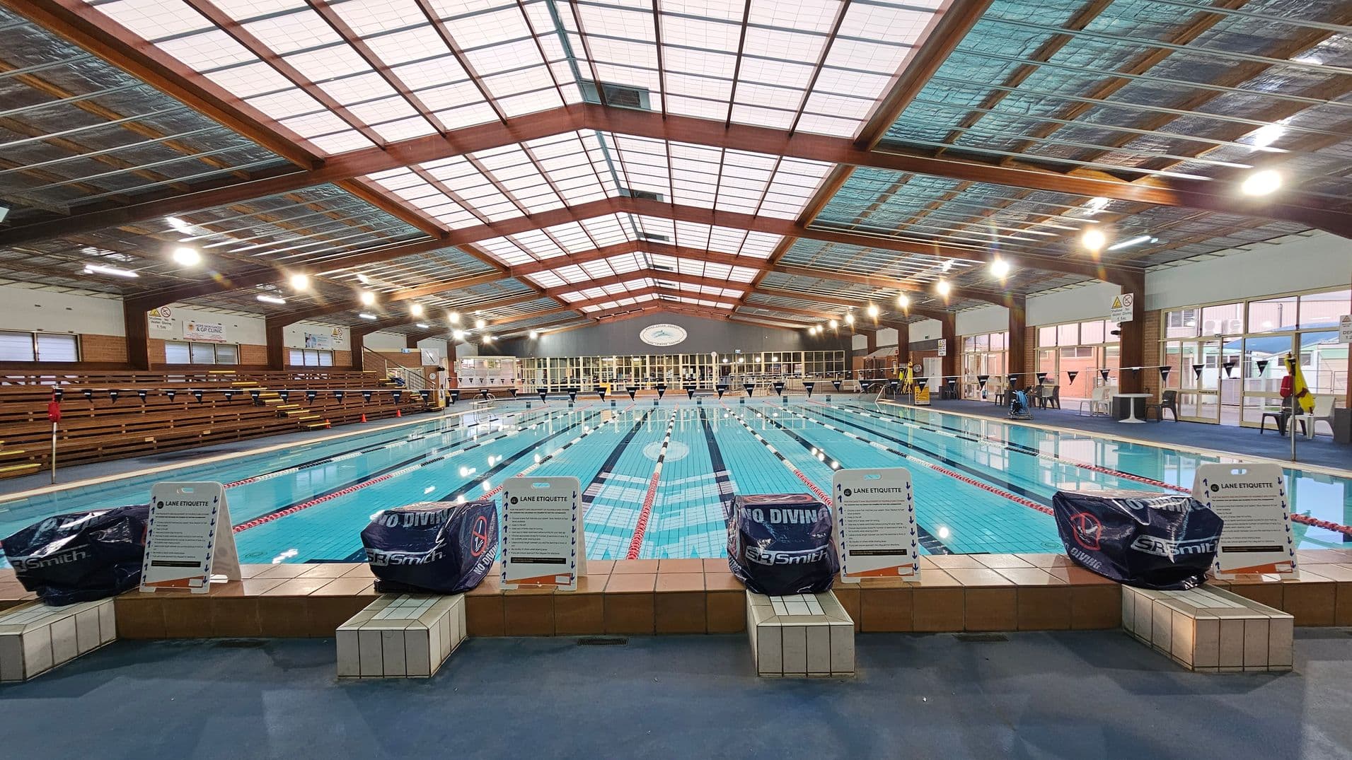 An indoor swimming pool with multiple lanes, surrounded by tiled flooring. On the left, there are wooden bleachers for spectators. The right side features large windows allowing natural light. The ceiling is high with a grid of lights and exposed beams. Signs with "Lane Etiquette" and "No Diving" are placed at the pool's edge. The pool is calm, with lane dividers visible.