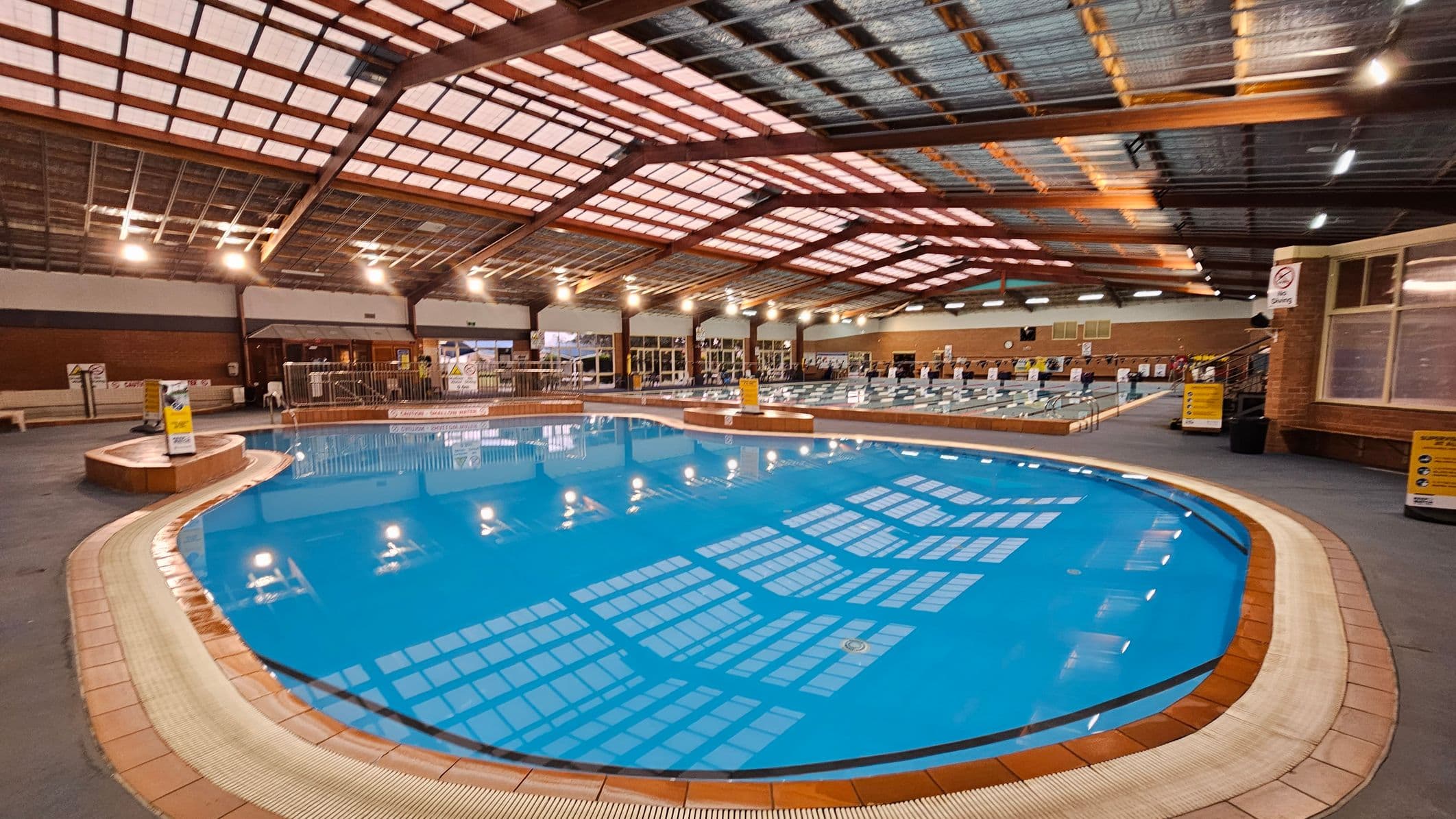 An indoor swimming pool with clear blue water is in the foreground, surrounded by a tiled edge. The ceiling is high with a grid of metal beams and translucent panels, allowing light to filter through. On the left, there are benches and a small fenced area. In the background, there are more pools and a row of large windows. The floor is grey, and there are various signs and notices around the pool area.