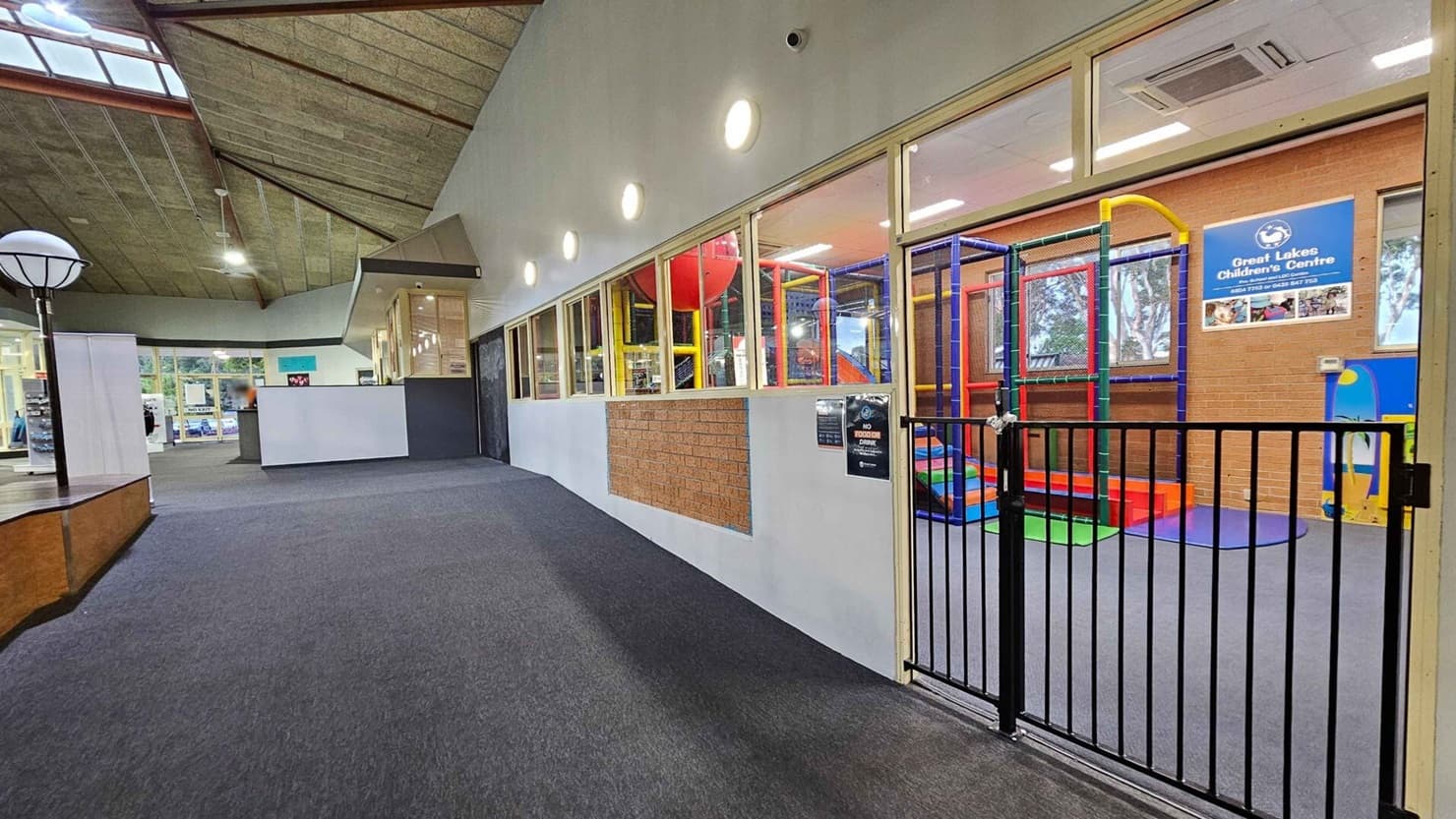 On the right, there's a children's play area with colourful climbing structures and a sign for the Great Lakes Children's Centre. A black gate separates the play area from the corridor. On the left, there are white walls and a reception desk. The ceiling is high with exposed beams and skylights, and the floor is carpeted in grey.
