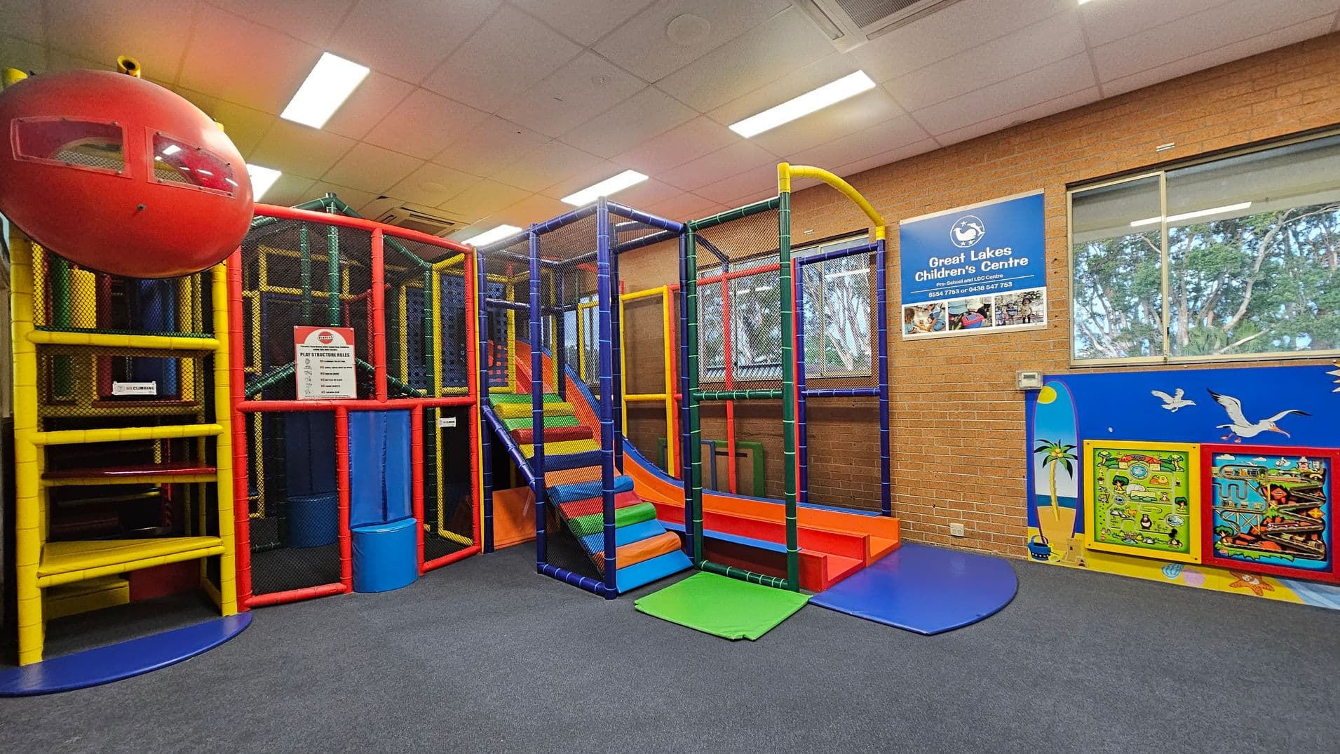 Indoor children's play area with a colourful climbing structure on the left, featuring red, yellow, and blue elements, and a red dome at the top. In the centre, a multi-coloured slide with steps. On the right, a brick wall with a window and a blue sign for "Great Lakes Children's Centre." Below the window, a mural with birds and palm trees, and two interactive wall panels. The floor is carpeted in grey, and the ceiling has fluorescent lights.