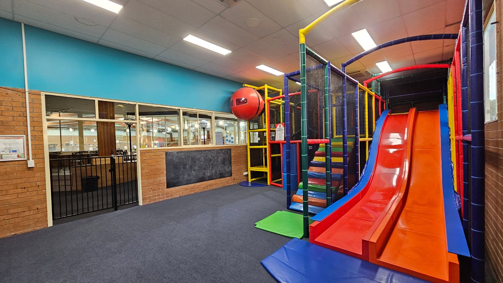 Indoor play area with a colourful slide on the right, featuring red and blue lanes. A climbing structure with yellow, red, and blue elements is next to it. On the left, a brick wall with a blackboard and a windowed partition overlooks another room. The floor is carpeted in dark blue, and the ceiling has white panels with fluorescent lights. A red spherical feature is attached to the climbing structure.